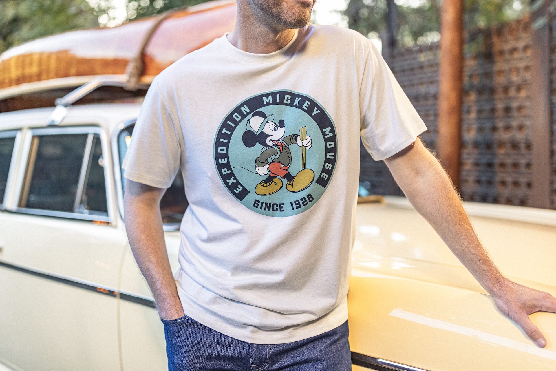 Man in a white Expedition Mickey Mouse t-shirt leans on a vintage cream car outdoors