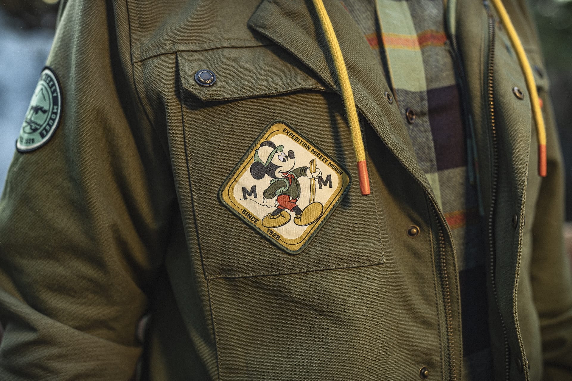 A green Expedition Mickey Mouse Collection jacket with a patch and yellow drawstrings, worn over a plaid shirt—park-ready style.