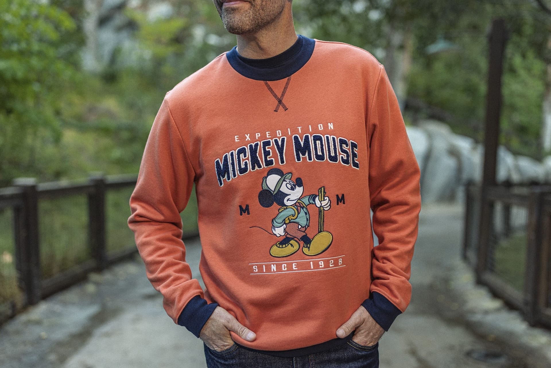 A man in an orange Mickey Mouse "Expedition" sweatshirt stands at Epic Universe, not far from the iconic castles of Disney Parks.