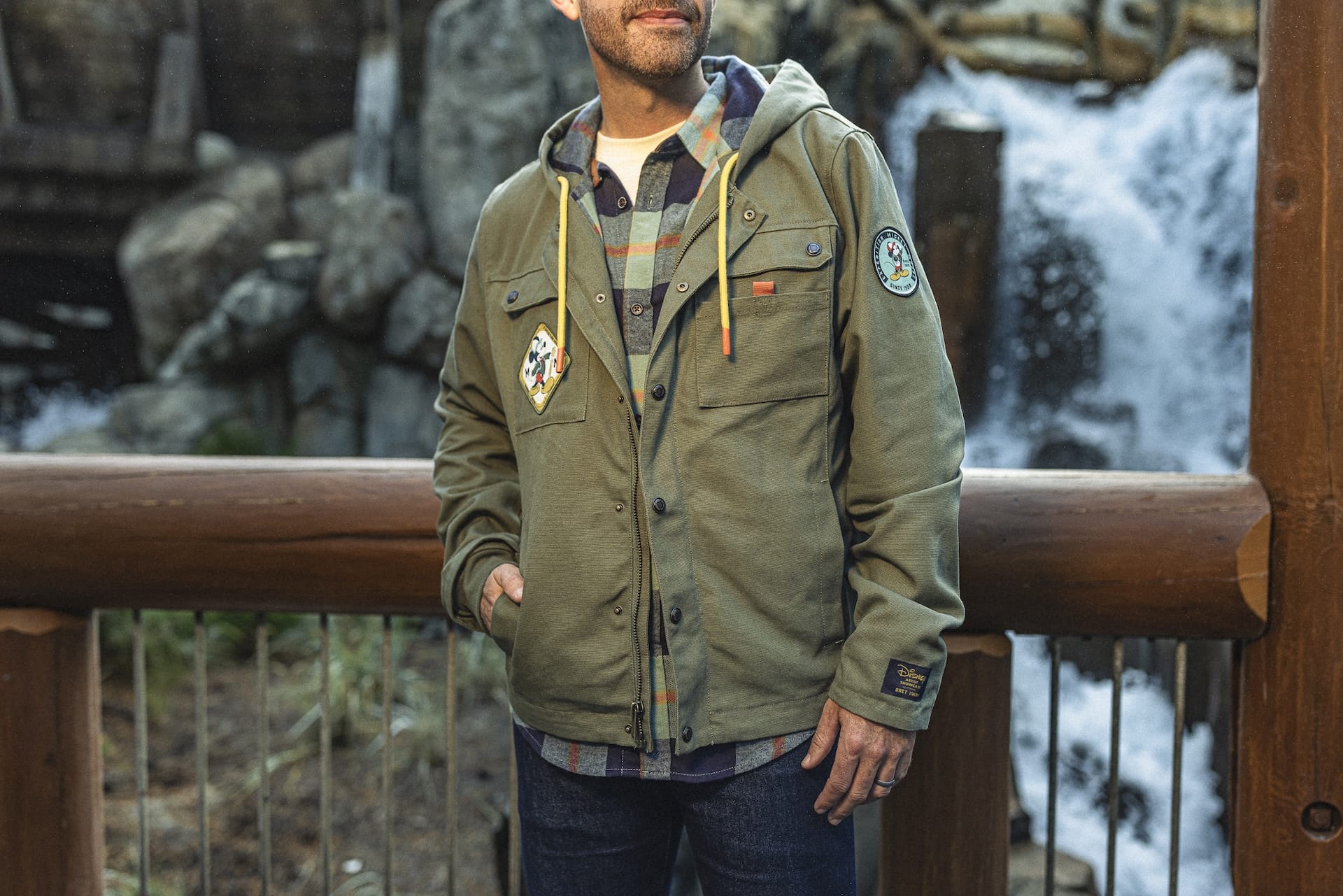 A man in a green hooded jacket and plaid shirt stands by a railing, ready for an Expedition Mickey Mouse