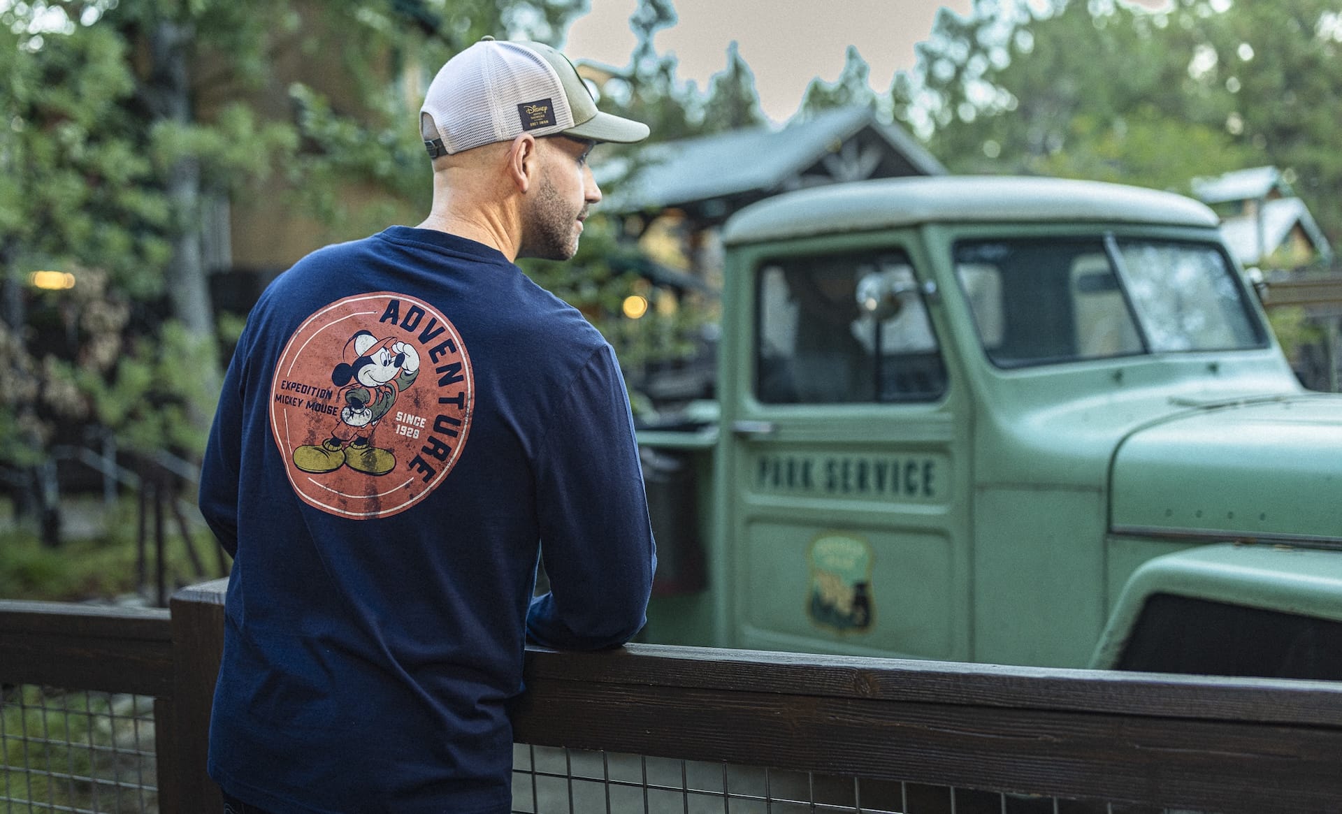 A man in a cap and Expedition Mickey Mouse Collection shirt stands by a fence, looking at a vintage green Park Service truck