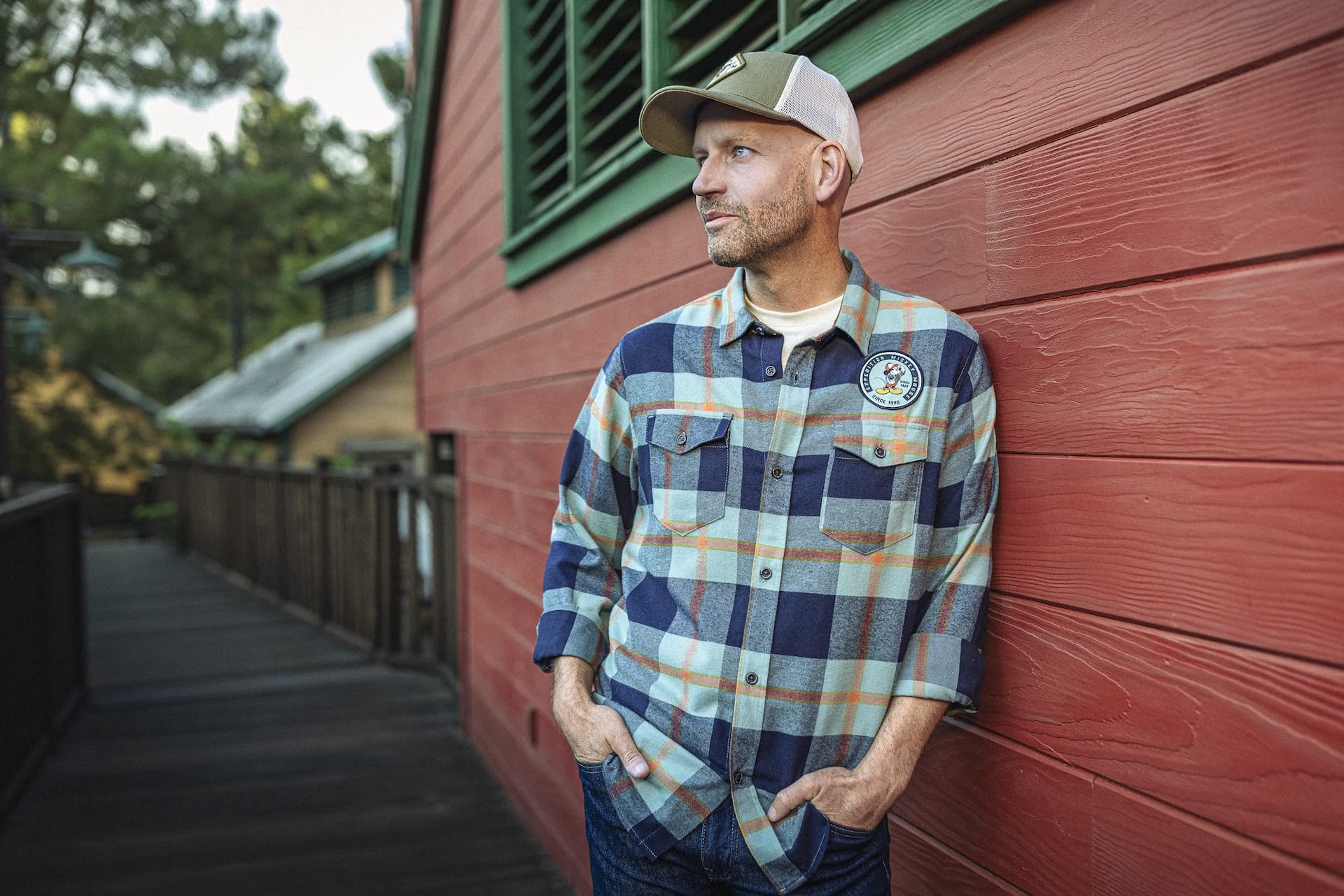 Man in Expedition Collection plaid shirt and cap leans on a red wall at Disney park, gazing left, hands in pockets.