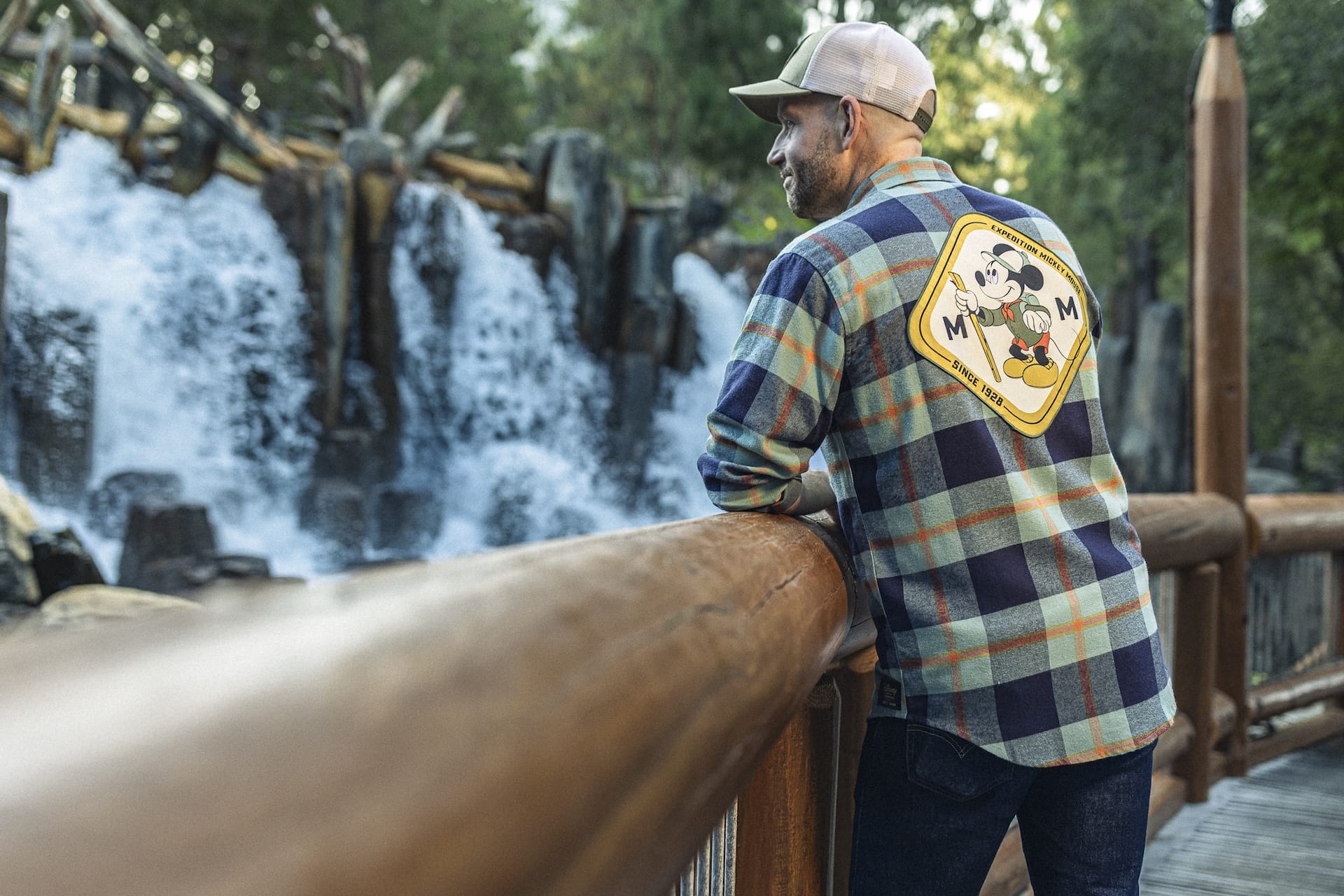 A man in a plaid shirt from the Expedition Mickey Mouse Collection stands by a railing, looking at a waterfall.