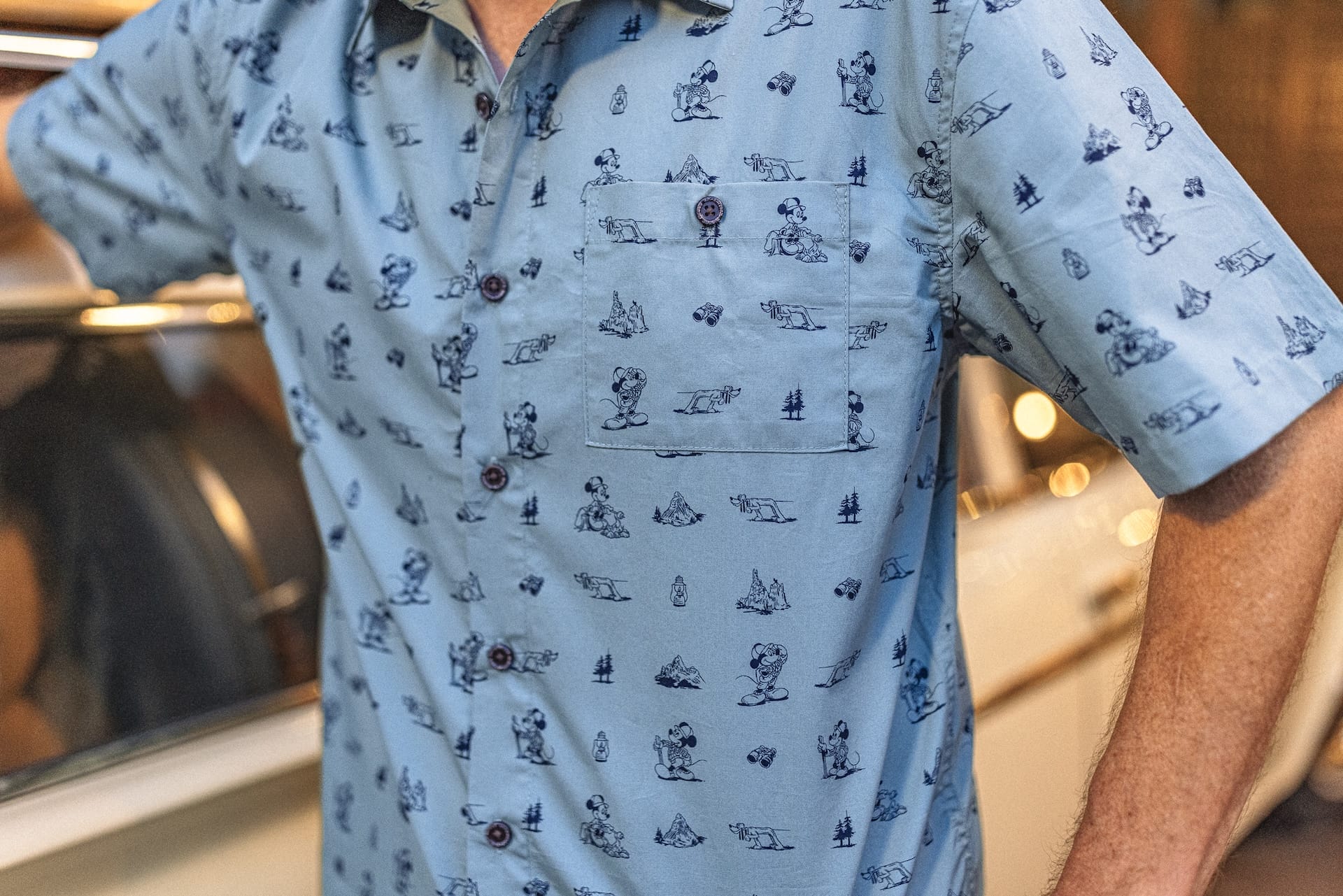 A guest in a light blue Expedition Mickey Mouse shirt with dog prints stands by a shiny surface.