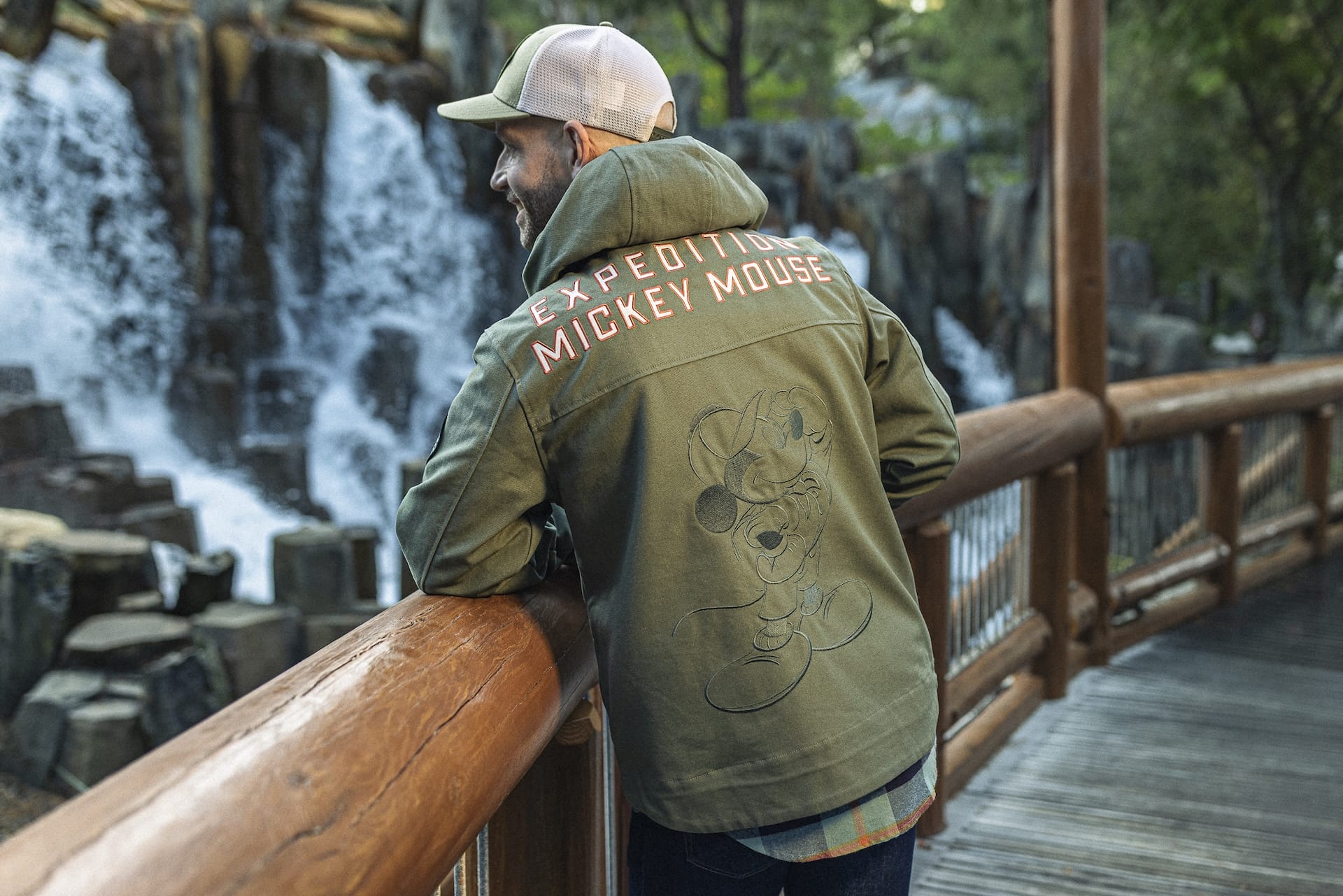 Man in a green Expedition Mickey Mouse jacket stands on a wooden bridge overlooking a park waterfall