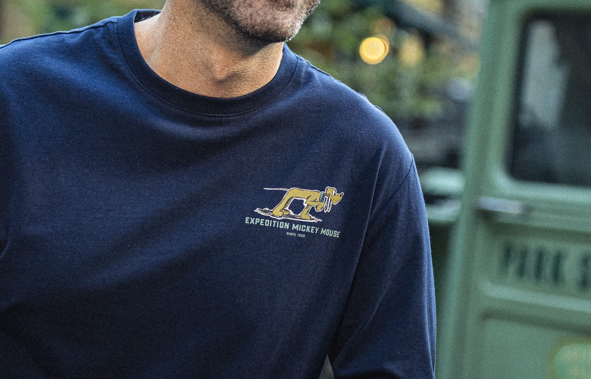 A visitor at a Disney park wears a navy Expedition Collection shirt featuring a yellow cheetah and "Expedition Mickey Mouse" logo.