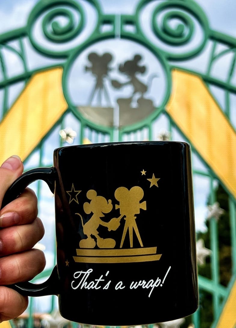 A hand holds a "That's a wrap!" Mickey Mouse mug near Walt Disney Studios Park’s entry gate—not a classic Disney park castle.