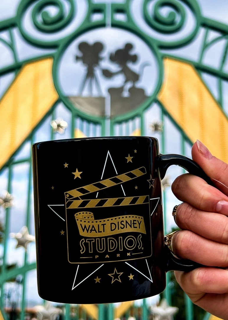 A hand holds a Walt Disney Studios Park Final Take Farewell mug in front of studio gates adorned with Mickey Mouse silhouettes, no castle.