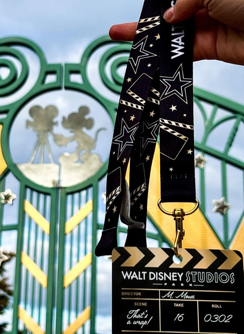 A guest displays a Walt Disney Studios Park Farewell lanyard badge in front of ornate gates topped with Mickey Mouse, unique to Paris.