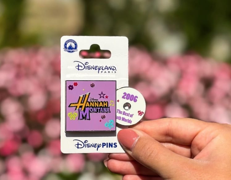A hand holds a Disneyland Paris Hannah Montana pin shaped like a CD jewel case.