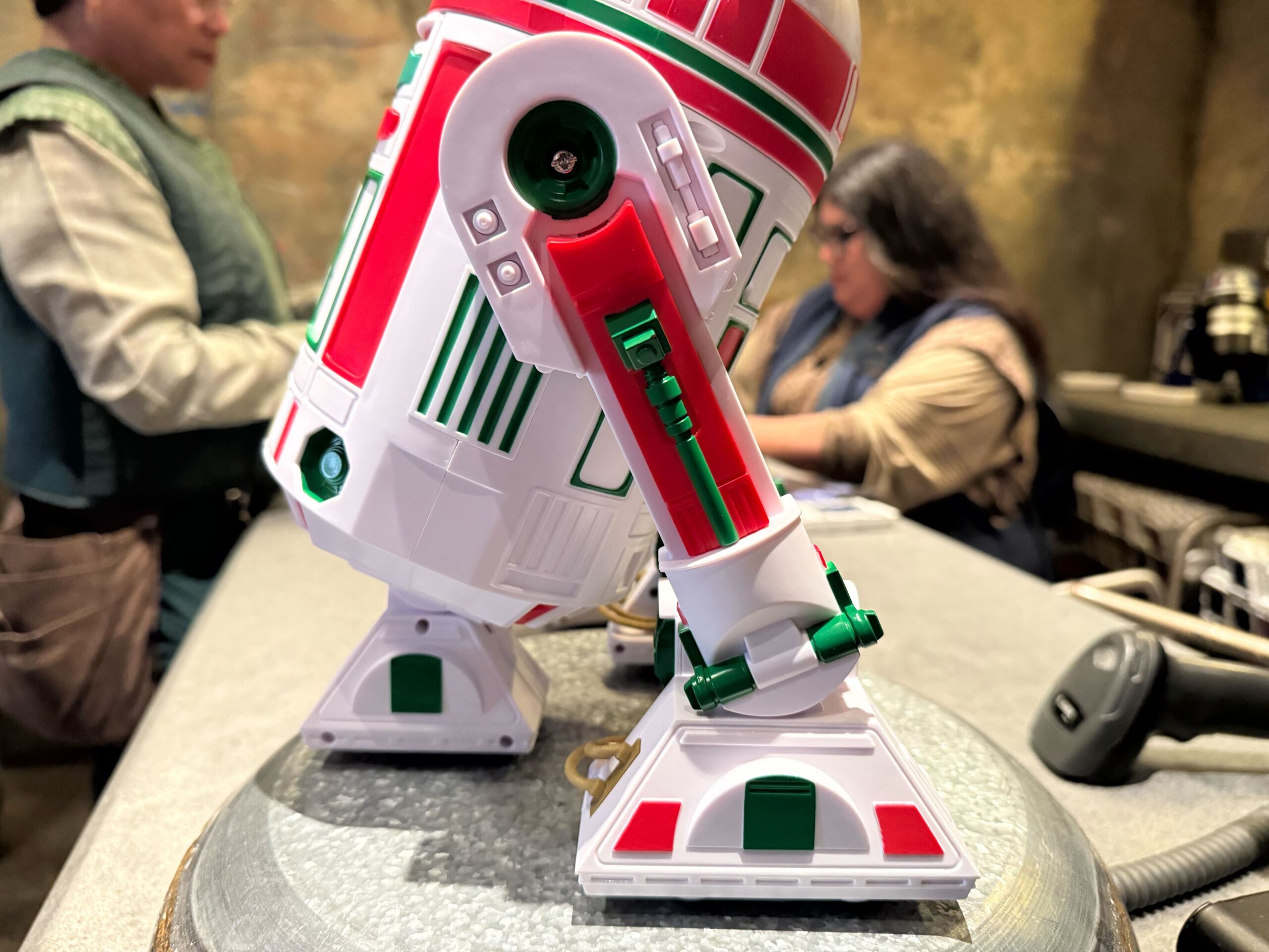 Close-up of a red, green, and white droid toy from the 2025 Disneyland merchandise on a counter, people blurred behind.
