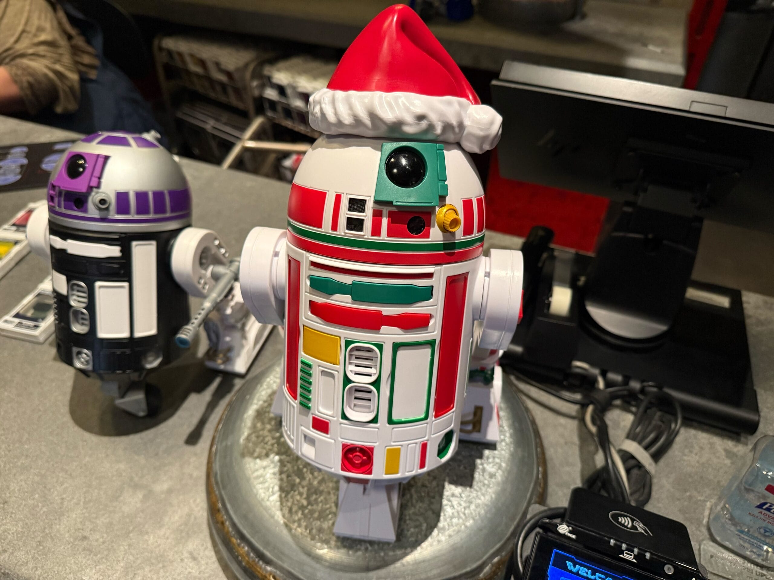 A colorful droid with a Santa hat sits by 2025 Disneyland Christmas merchandise near a register, next to another festive droid.