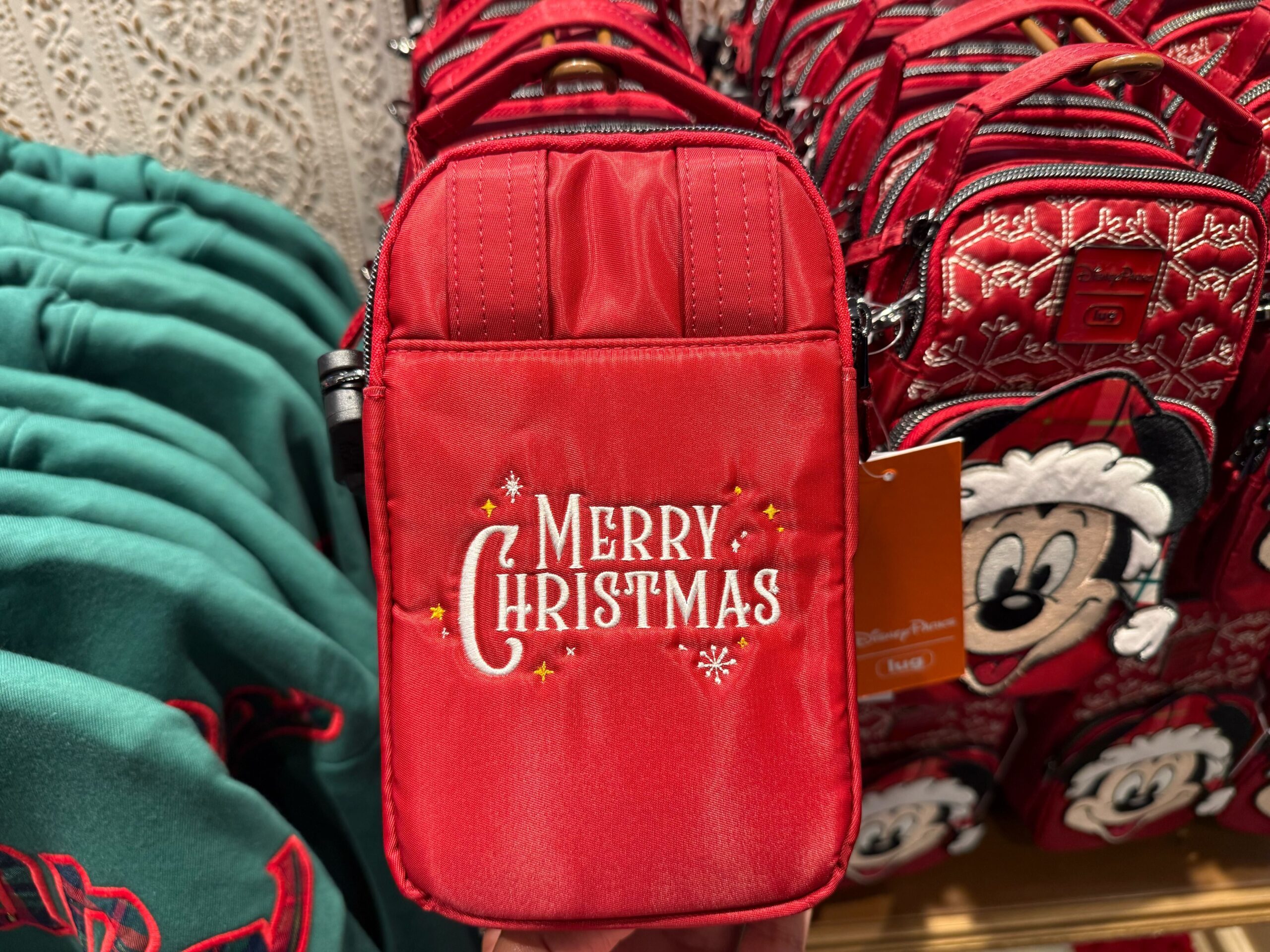 A red pouch with "Merry Christmas" embroidered, displayed among 2025 Disneyland Christmas merchandise and holiday bags.