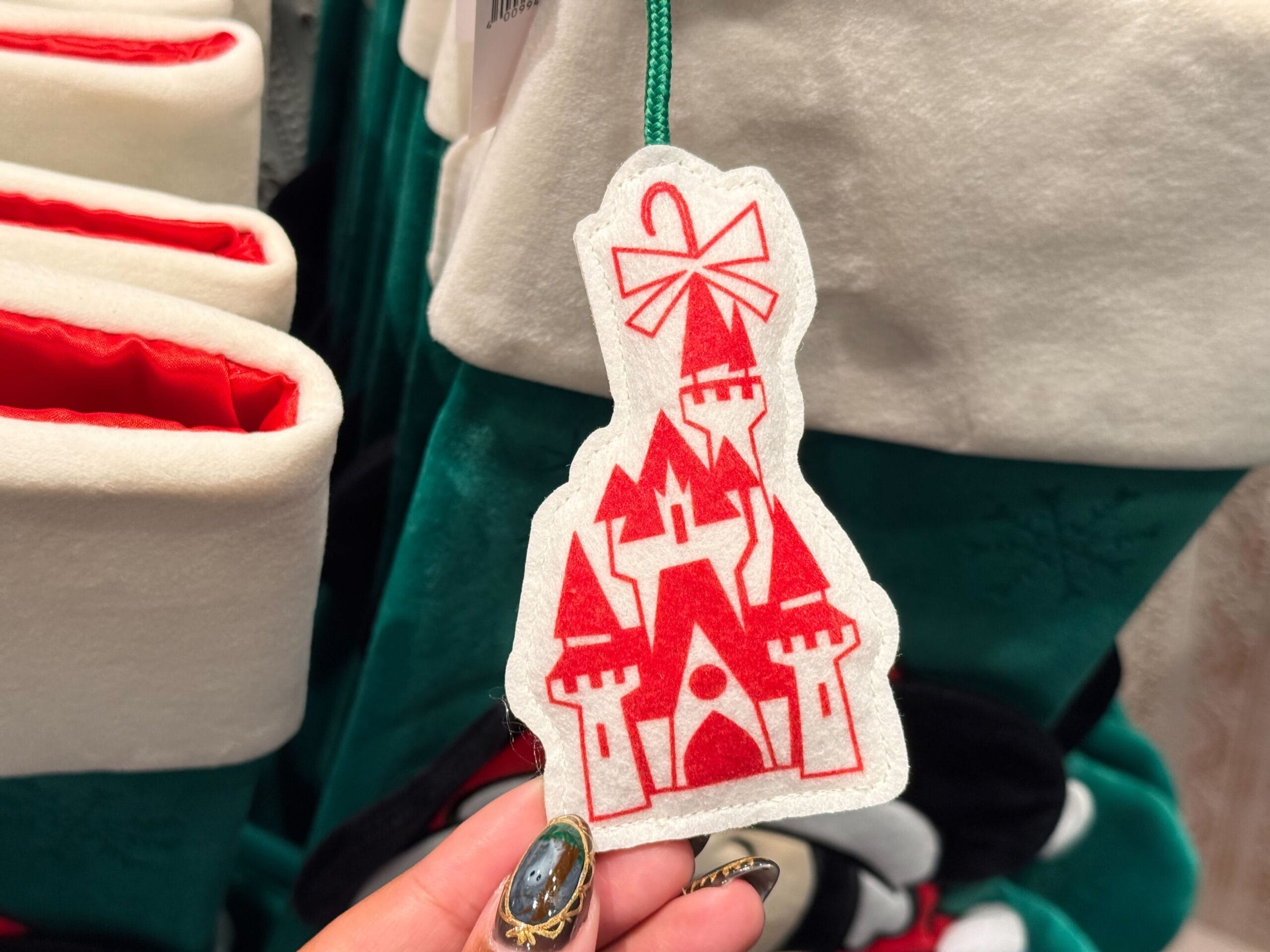 A hand holds a white felt ornament with a red castle, part of Disneyland Christmas Merchandise 2025, near stockings.