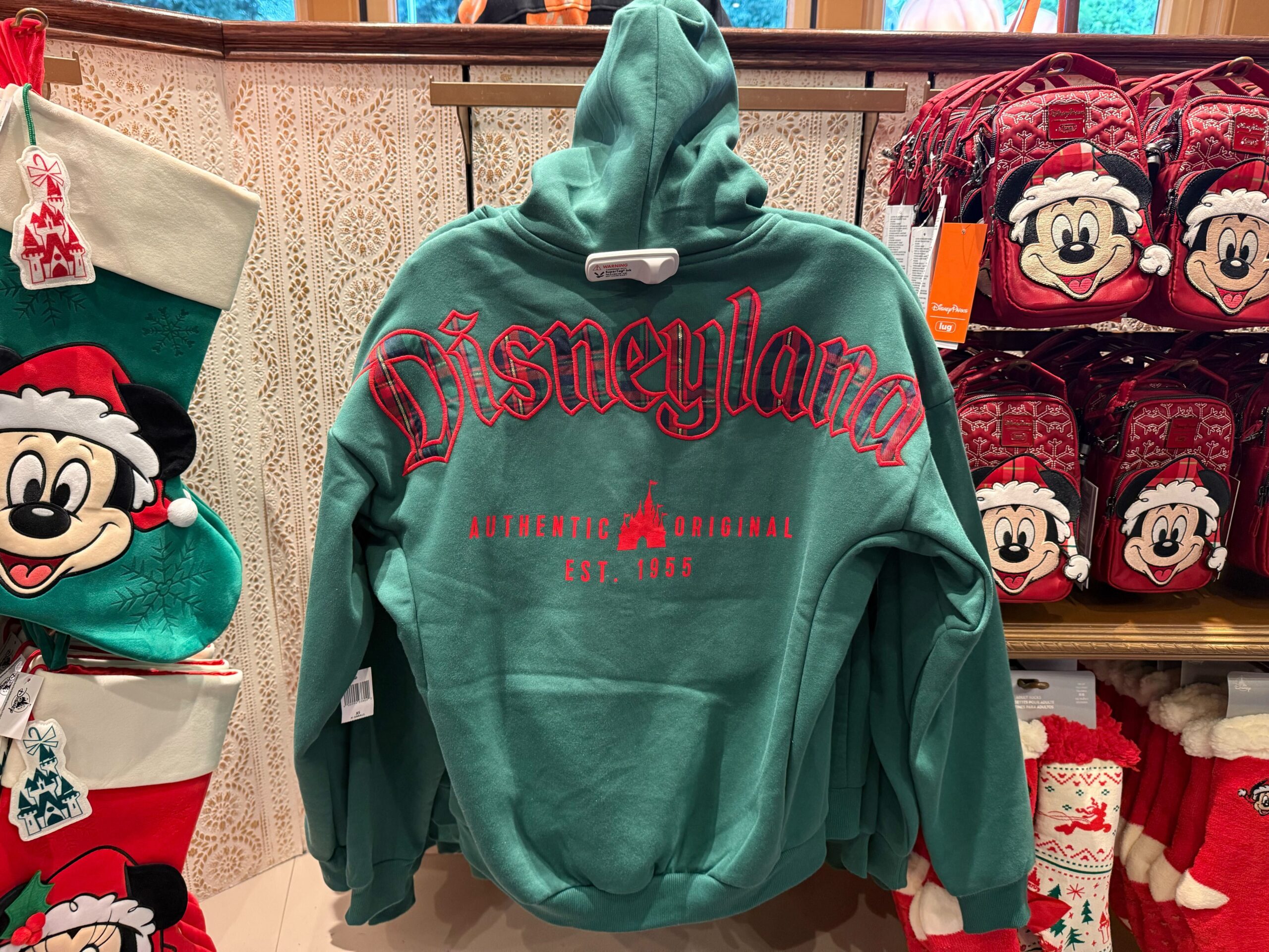 Green hoodie with red "Disneyland" text among Disneyland Christmas merchandise, holiday Mickeys, and 2025 prices on display.
