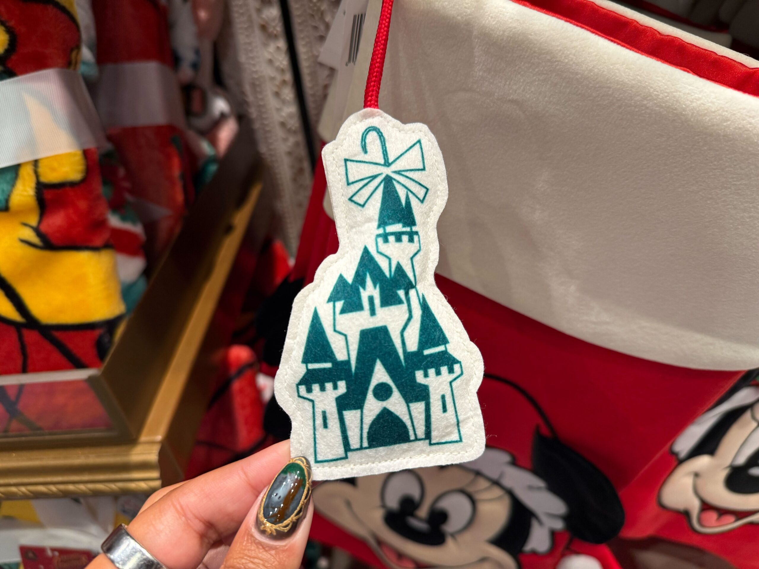 Hand holding a felt teal castle ornament, part of 2025 Disneyland Christmas Merchandise, near a holiday stocking.
