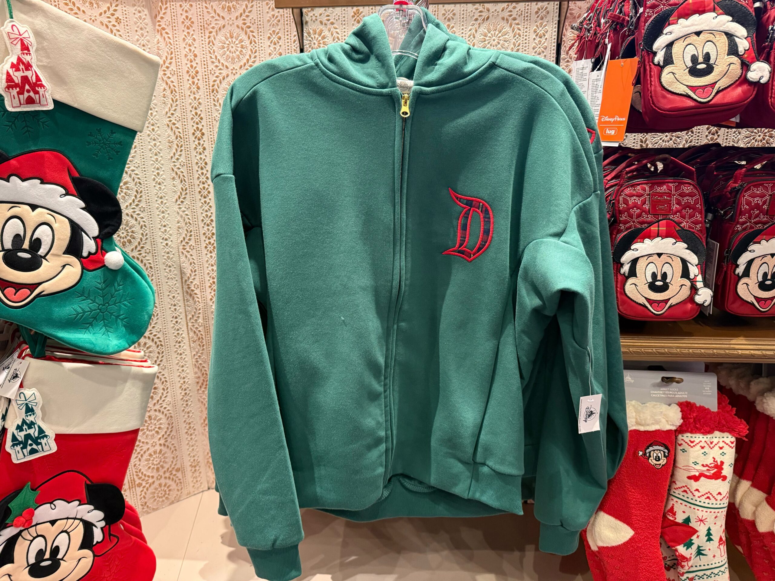 Green zip-up jacket with a red "D" logo, among 2025 Disneyland Christmas merchandise and Mickey Mouse stockings.