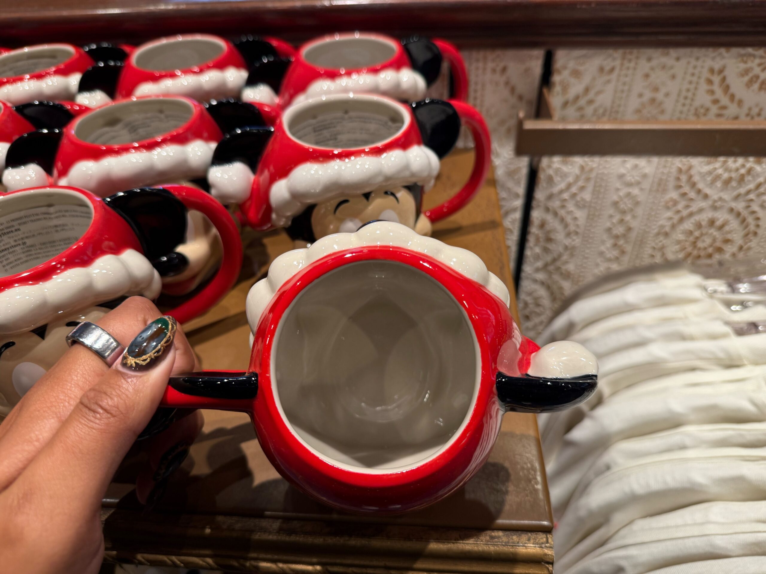 A hand holds a red mug shaped like Minnie Mouse’s head, part of 2025 Disneyland Christmas merchandise on the shelf.