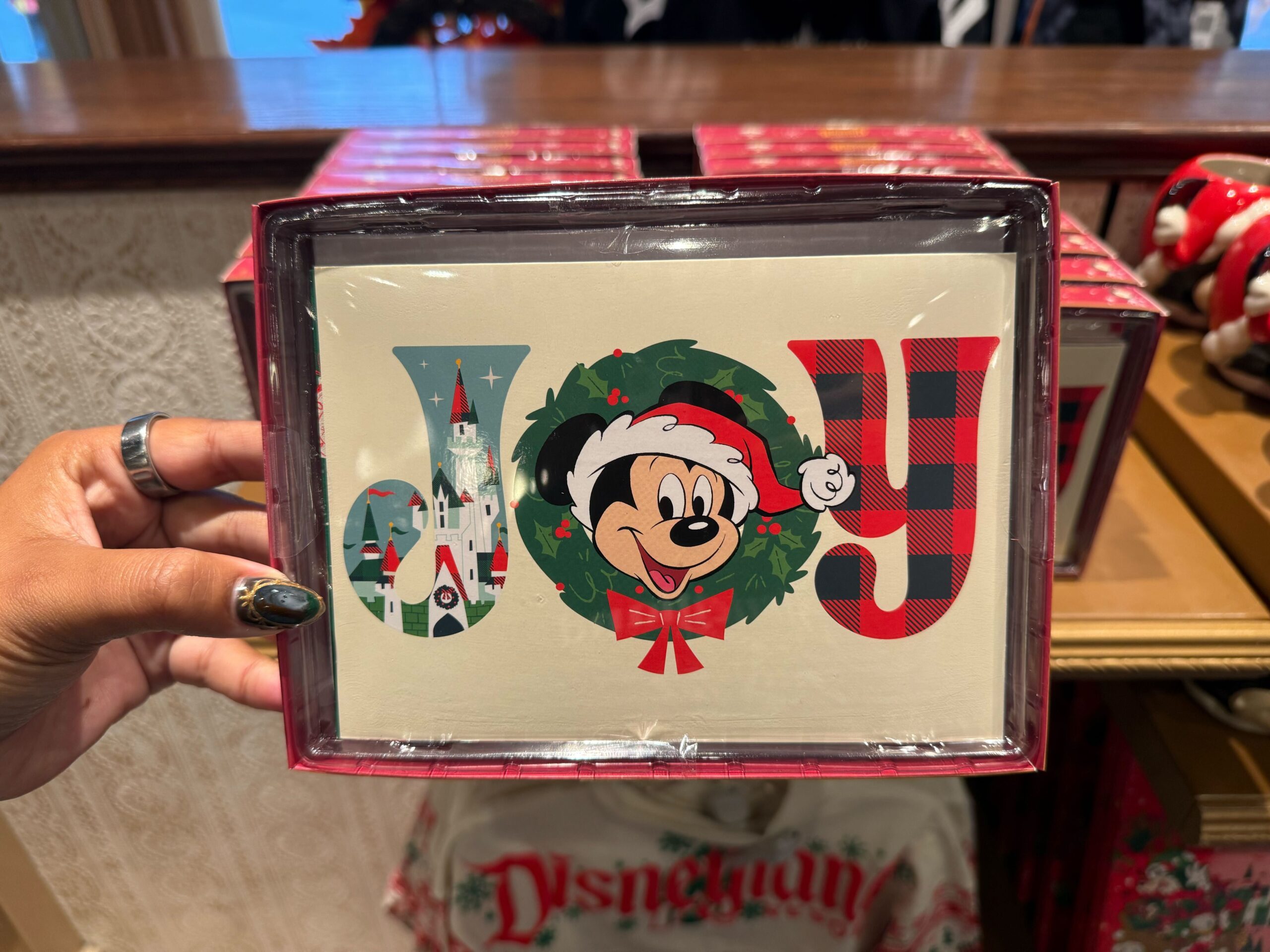 Hand holds a 2025 Disneyland Christmas merchandise card reading "JOY," featuring Mickey, a castle, and plaid details.