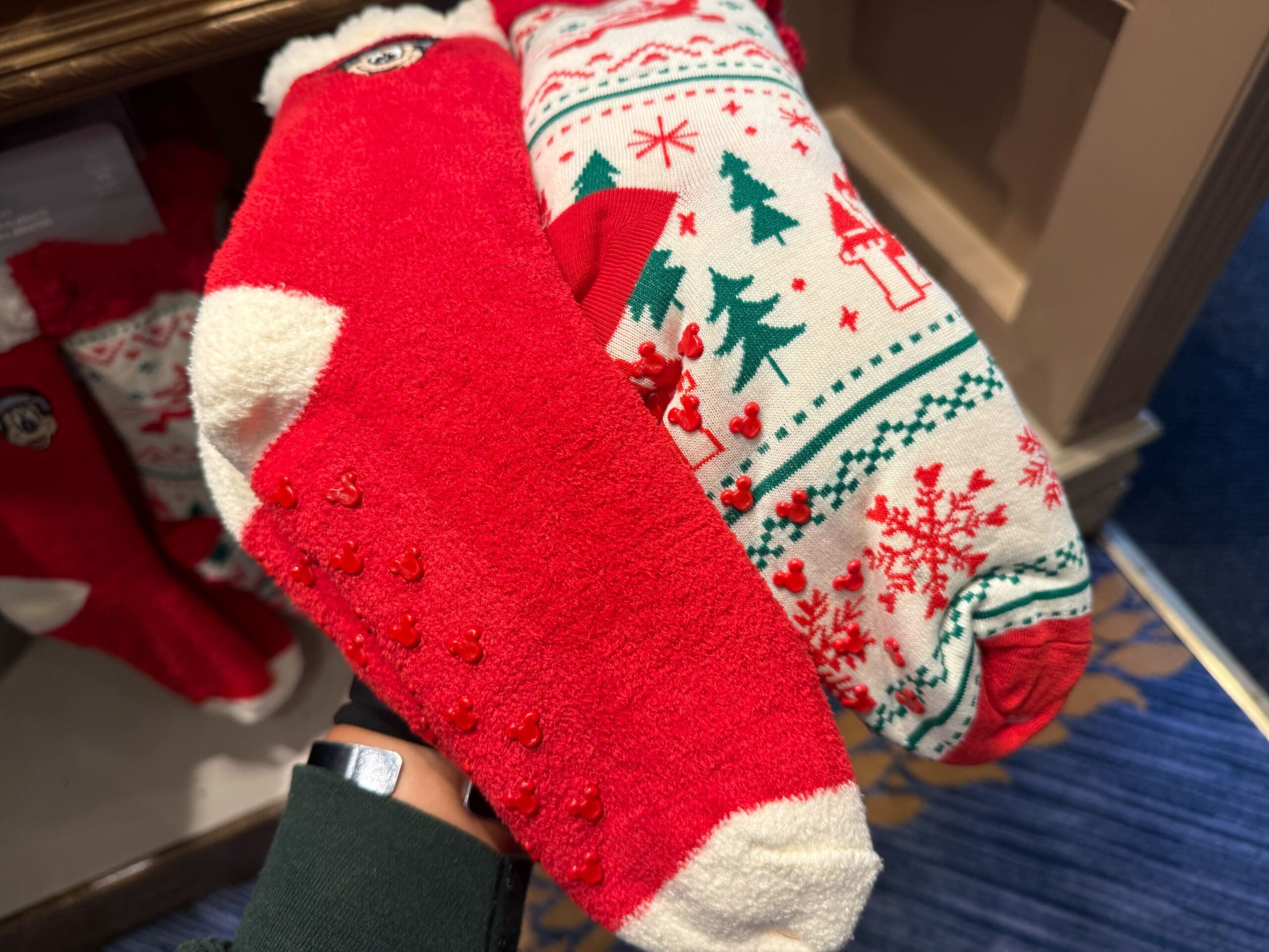 A hand holds two fuzzy holiday socks—2025 Disneyland Christmas merchandise with festive patterns and grip dots.