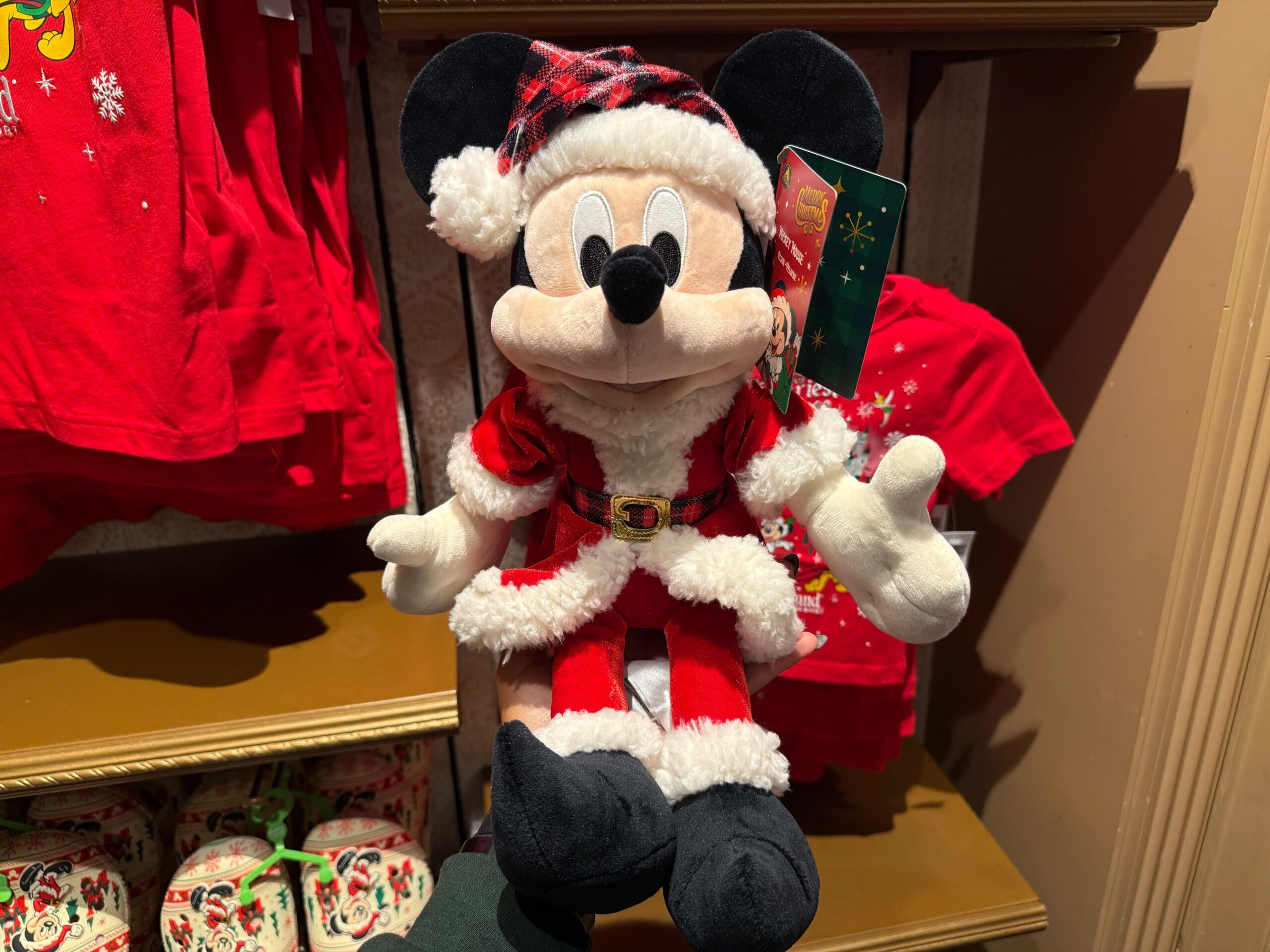 Plush Mickey Mouse in a Santa outfit, part of 2025 Disneyland Christmas merchandise, displayed with red shirts behind.