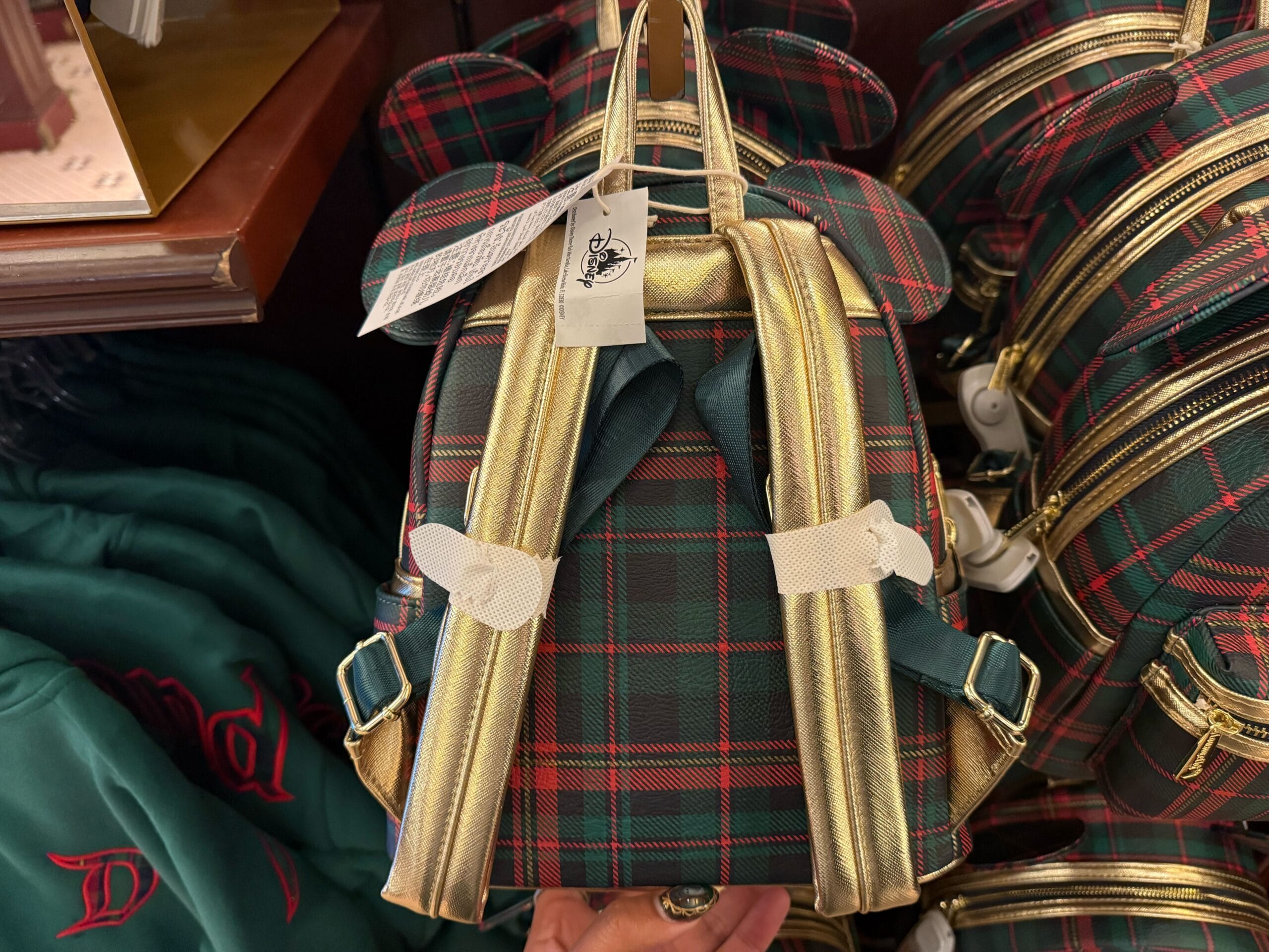A Disneyland Plaid Christmas Loungefly mini backpack with gold straps and mouse ears hangs in a park shop, echoing festive park style.