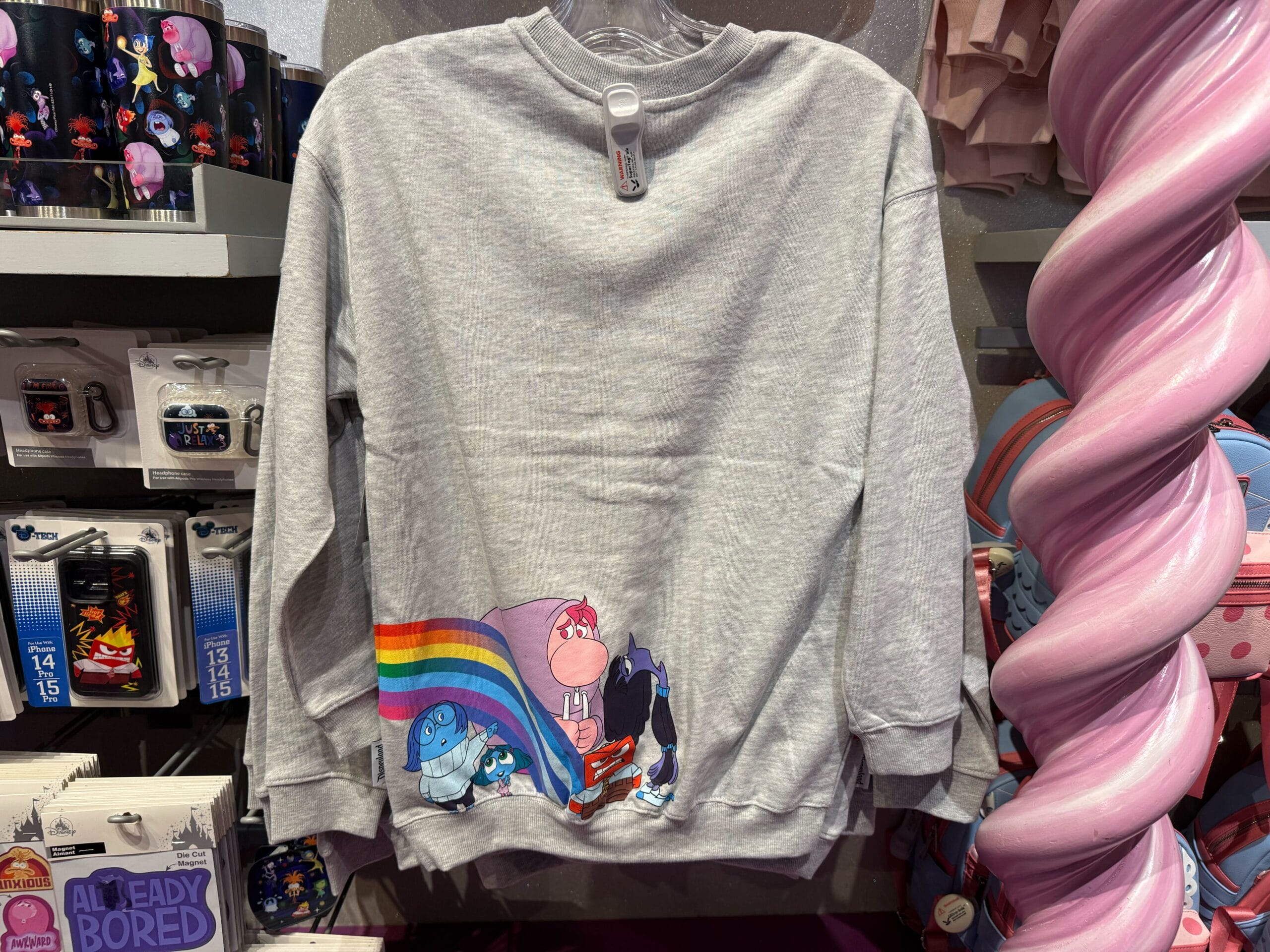 Gray sweatshirt featuring Disney cartoon characters, a rainbow, and clouds, displayed near mugs and pastel decor in a Disney Park shop.