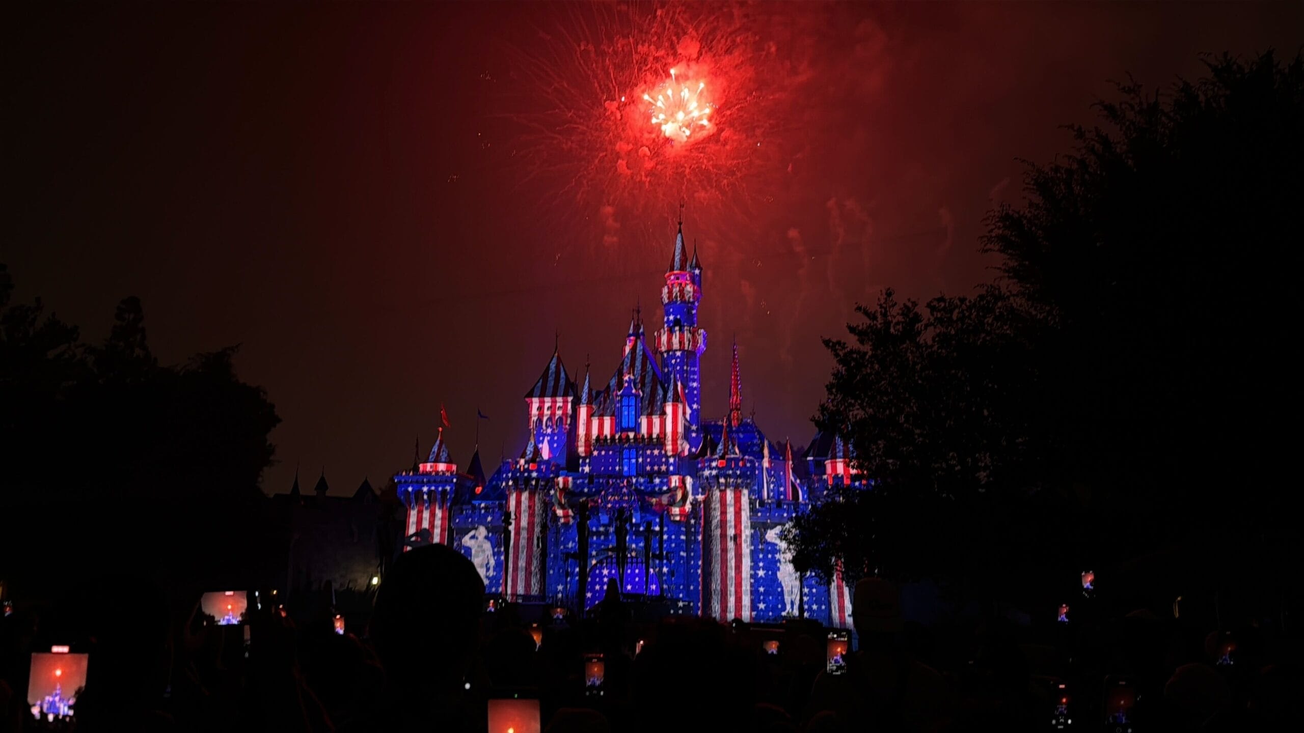 Disneyland castle, unlike the taller Cinderella Castle in Walt Disney World, glows in red, white, and blue as fireworks light up the night.