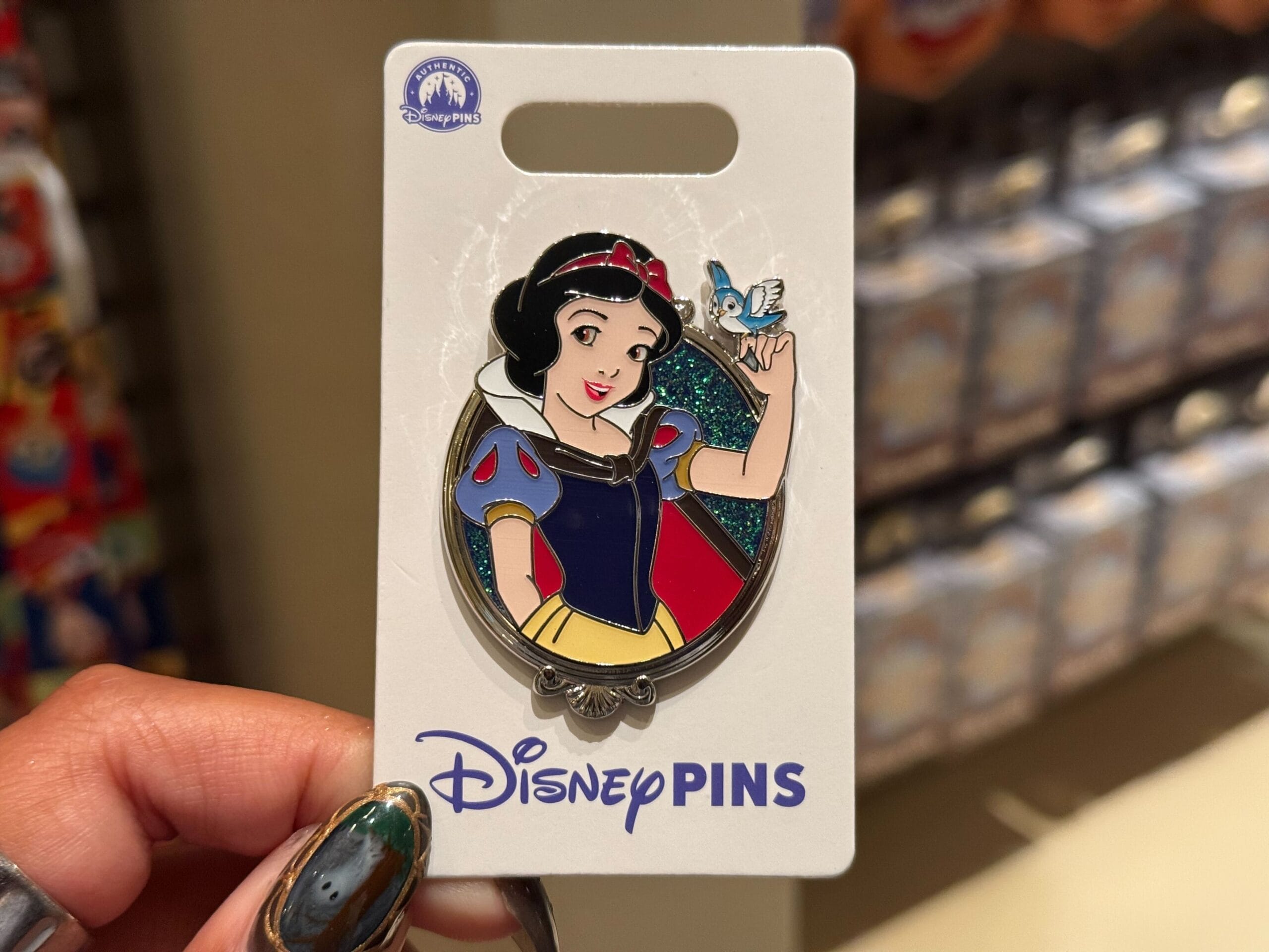 A hand holds a Princess Pins Disney pin of Snow White smiling with a bluebird, shown on a Disney card sold at Disney Parks worldwide.