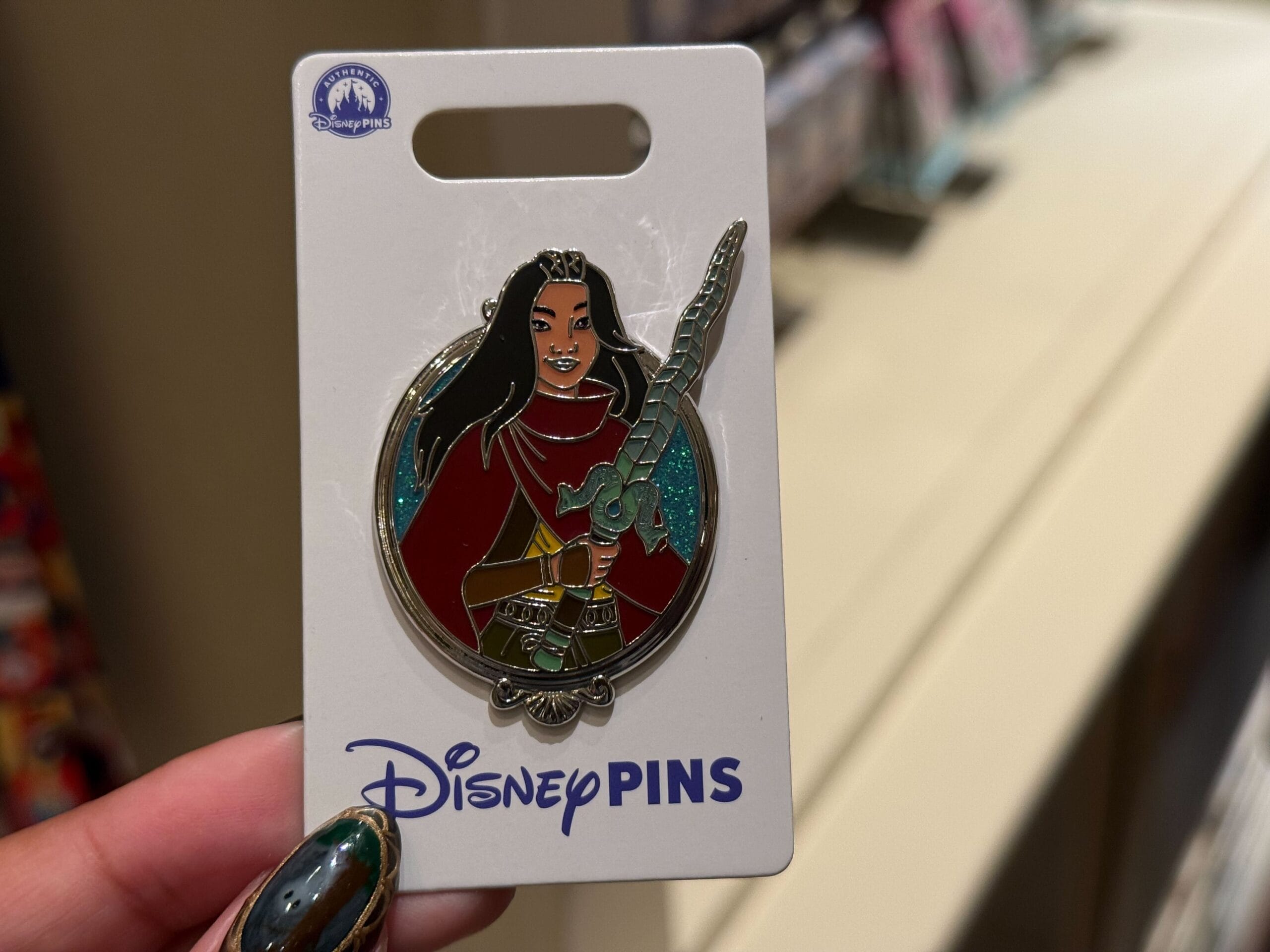 A hand holds a Disney Princess pin of Mulan in armor with a sword, on a “Disney Pins” card, typical of souvenirs at Disney parks.