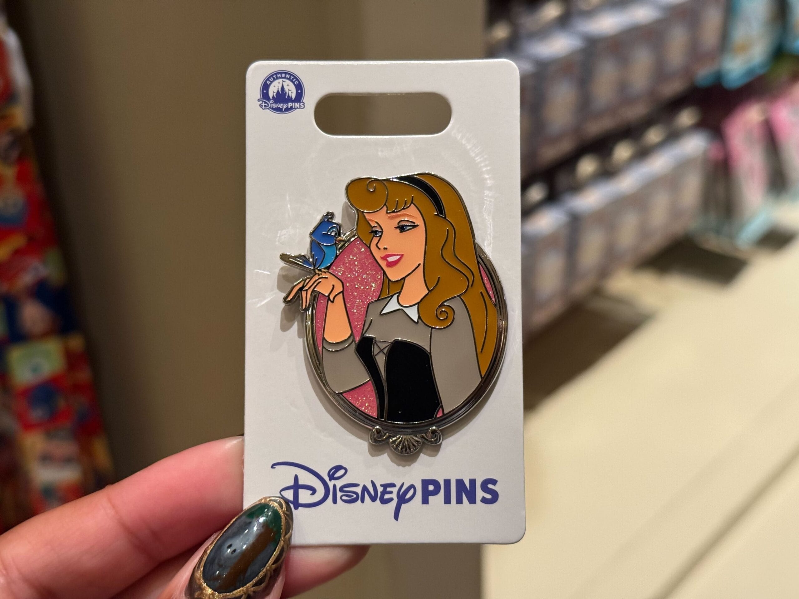 A hand holds a Disney Princess Pins card showing Princess Aurora with a bluebird, displayed in a Disney parks gift shop.