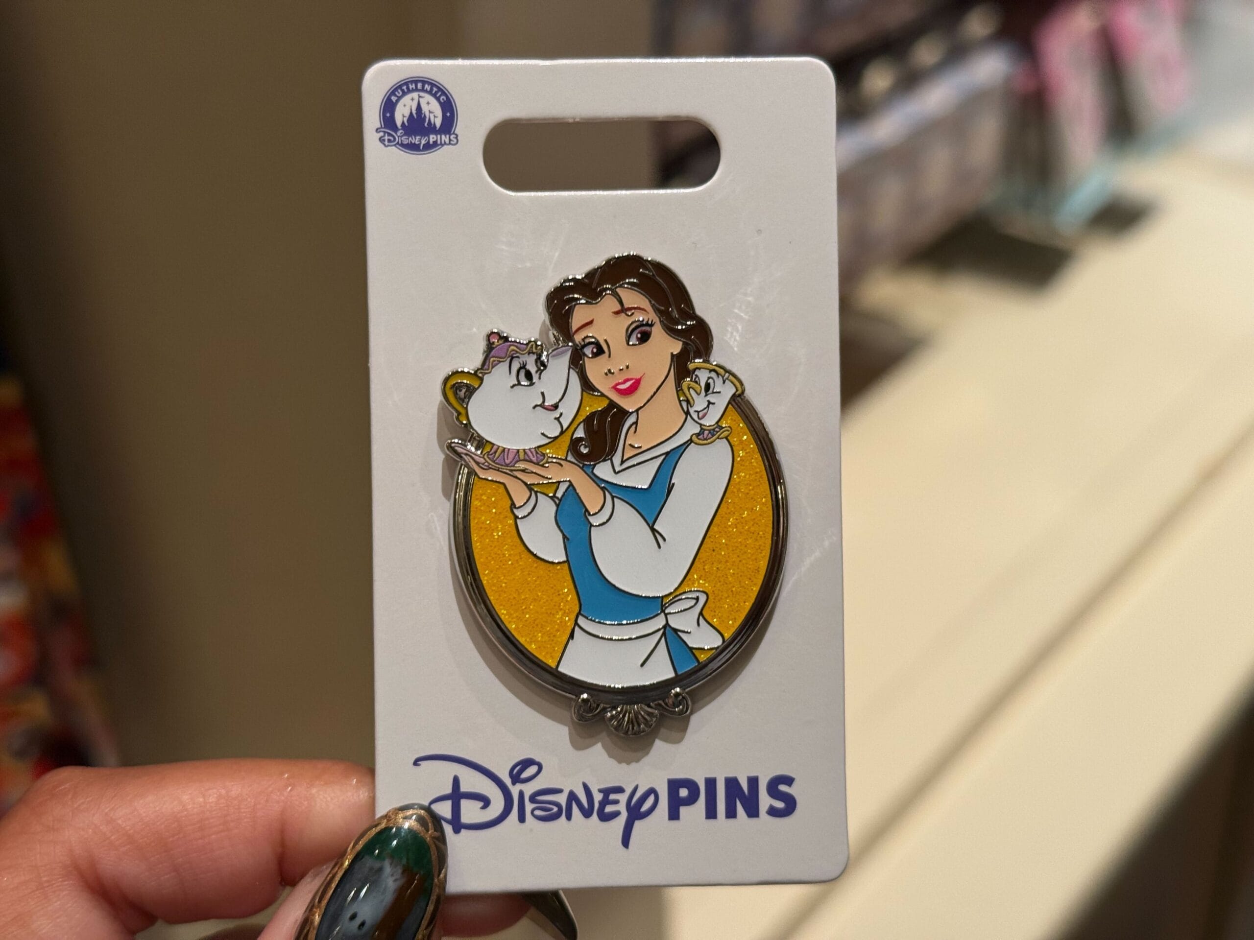 A hand holds a Belle pin with Mrs. Potts and Chip from "Beauty and the Beast," similar to pins found at Disney Parks worldwide.