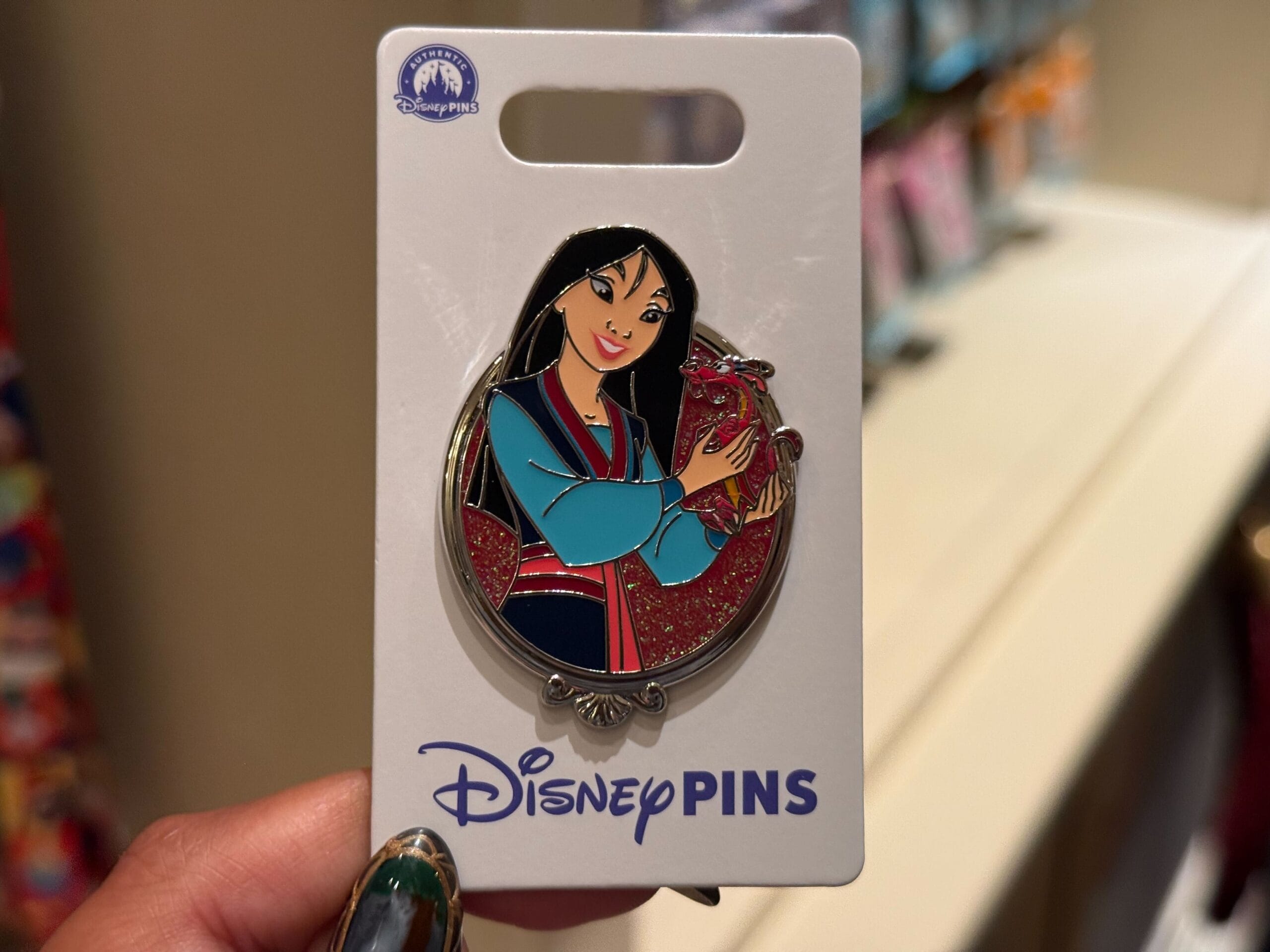 A Princess Pins Disney pin of Mulan holding Mushu, displayed on a backing card and held by hand, sold at Disney parks worldwide.