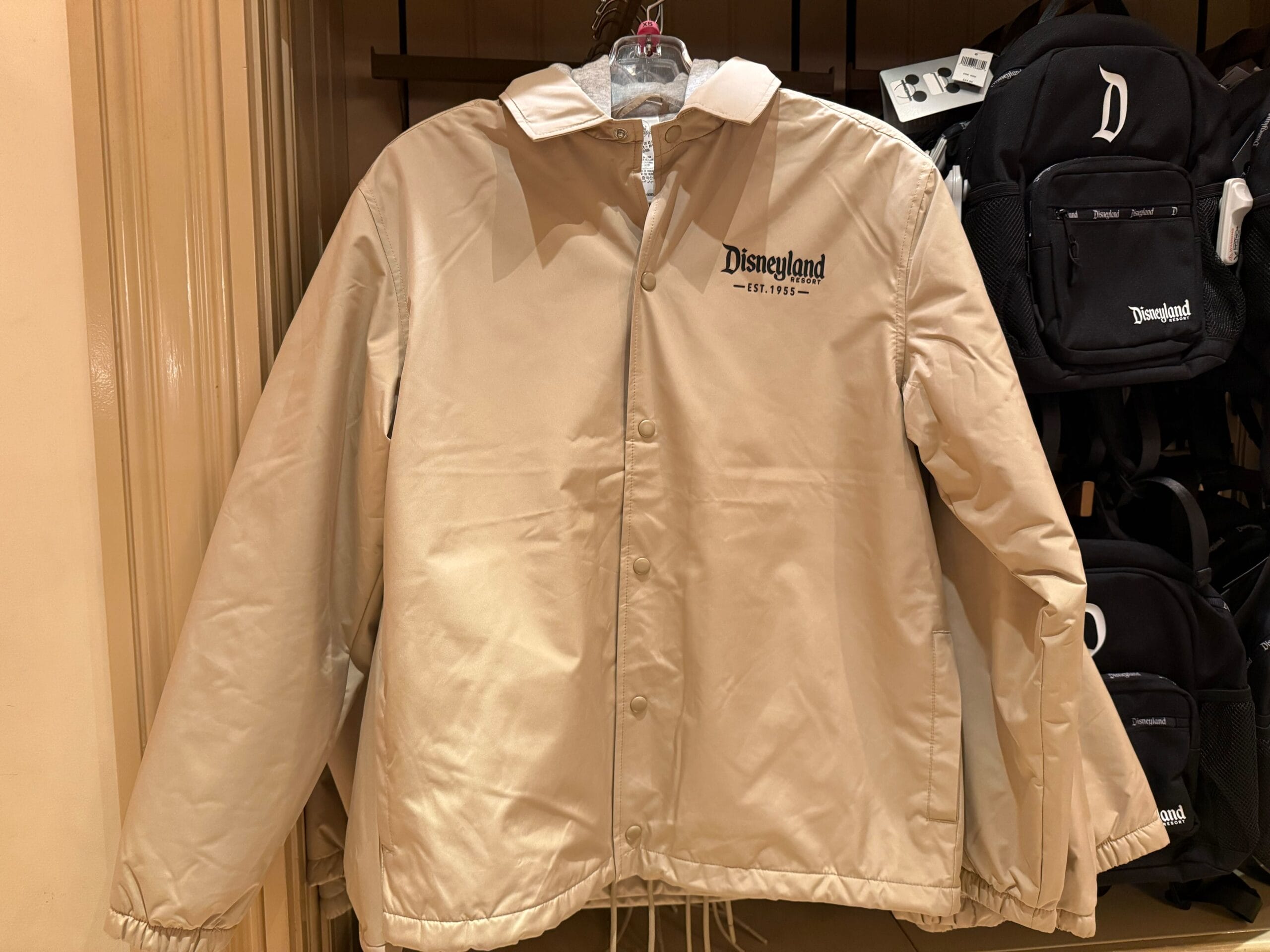 Tan Disneyland jacket with a white collar on a hanger, next to black Disneyland backpacks in a park shop, no castle in view.