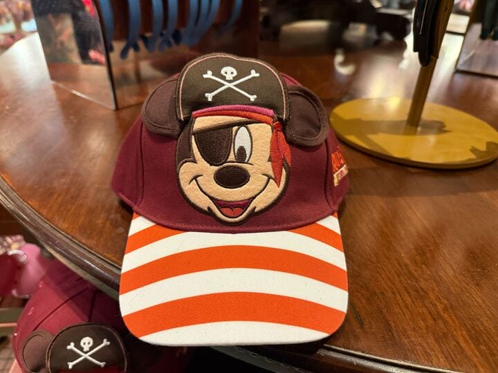 A maroon pirate hat featuring Mickey Mouse’s face, classic Disneyland merchandise, rests on a wooden table near park souvenirs.