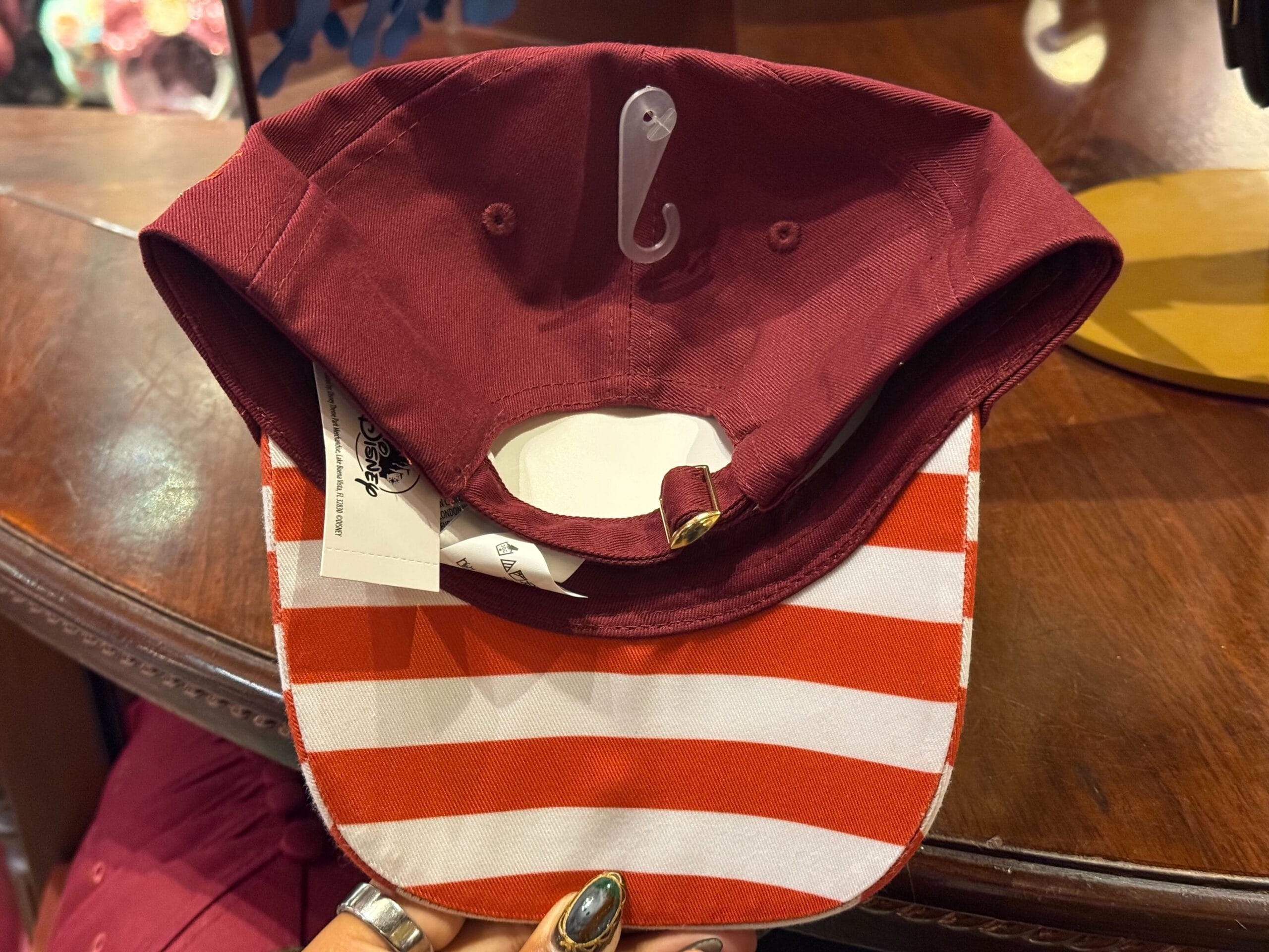 A maroon cap with a red and white striped bill, inspired by Disneyland merch, features a white fish hook embroidered inside the brim.