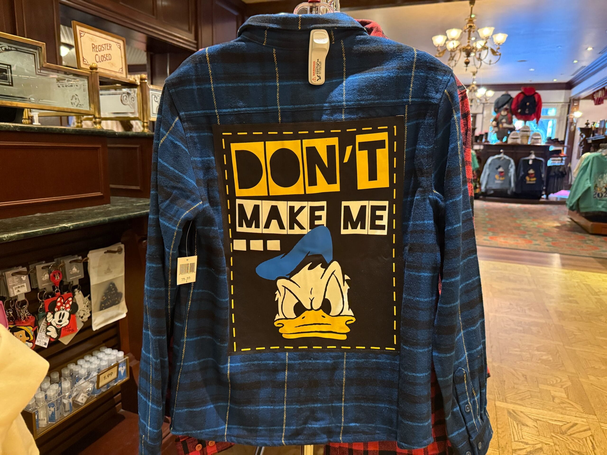A blue plaid shirt featuring Donald Duck and the phrase "DON'T MAKE ME," spotted among playful Disneyland park merchandise displays.
