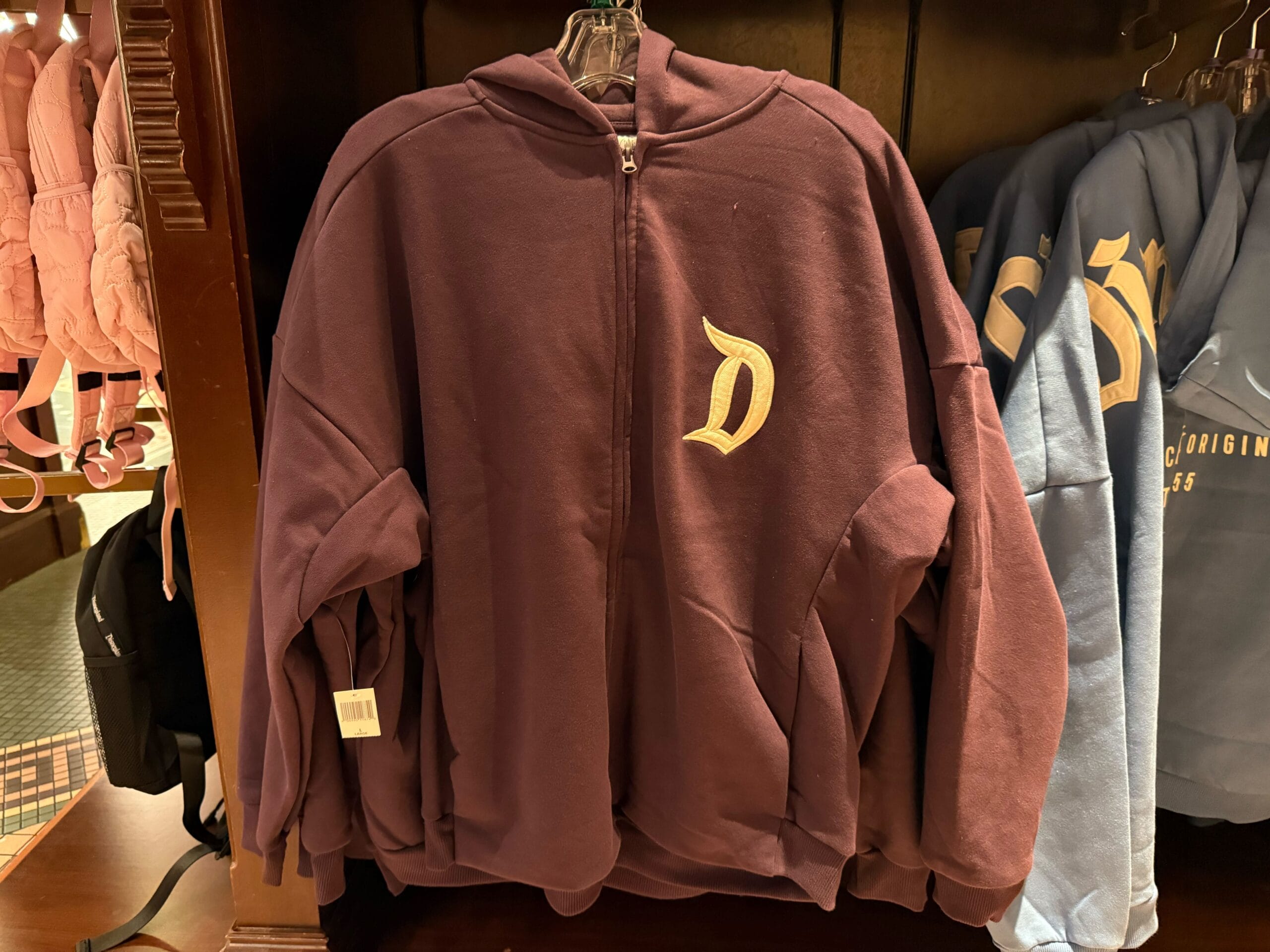 A maroon zip-up hoodie with a gold "D" on the chest, ideal as a Disneyland gift, hangs on a rack near park merchandise.