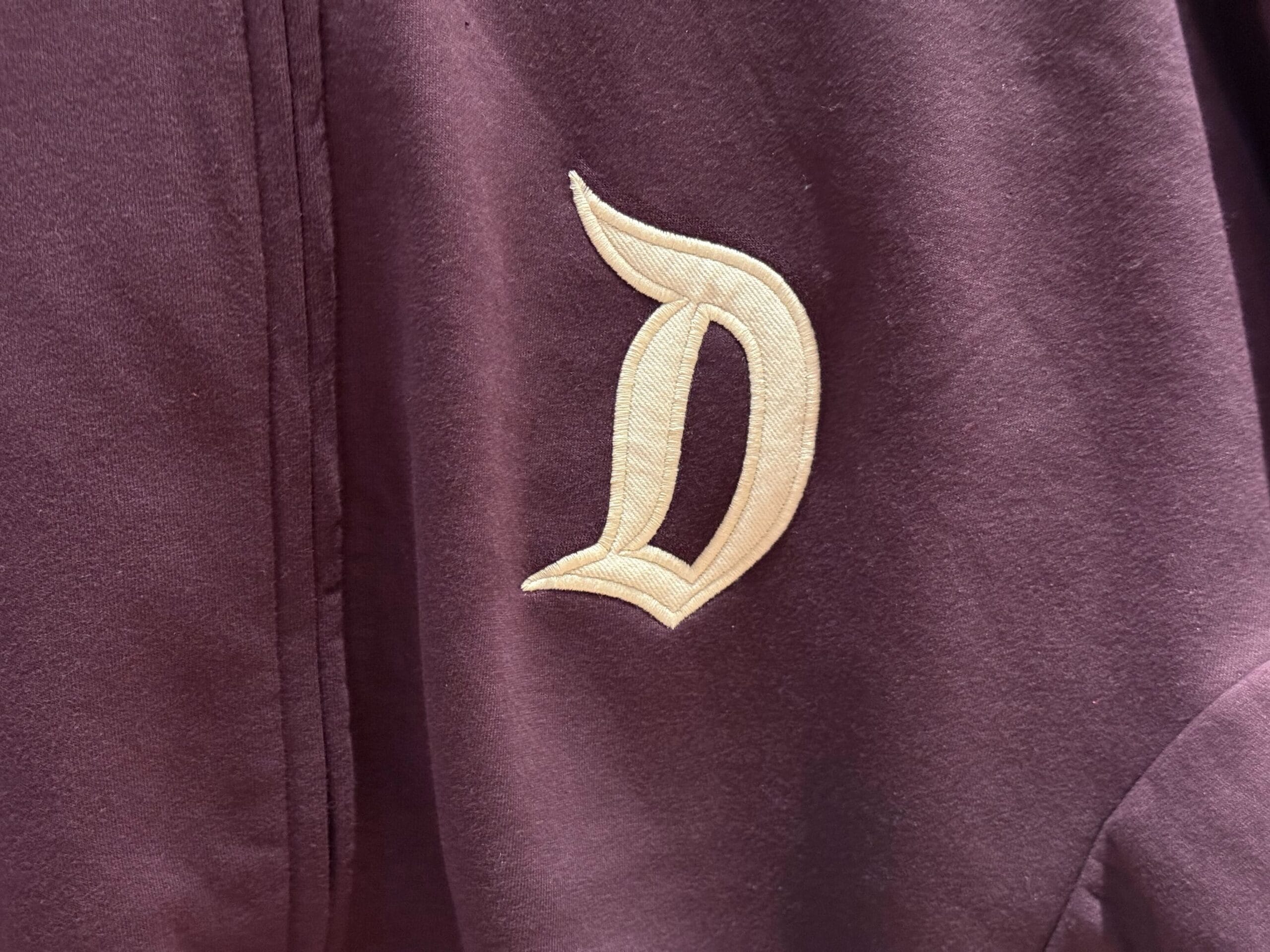 A cream-colored letter "D" embroidered on maroon fabric, echoing classic Disneyland logos and ideal for Disney park collectors.
