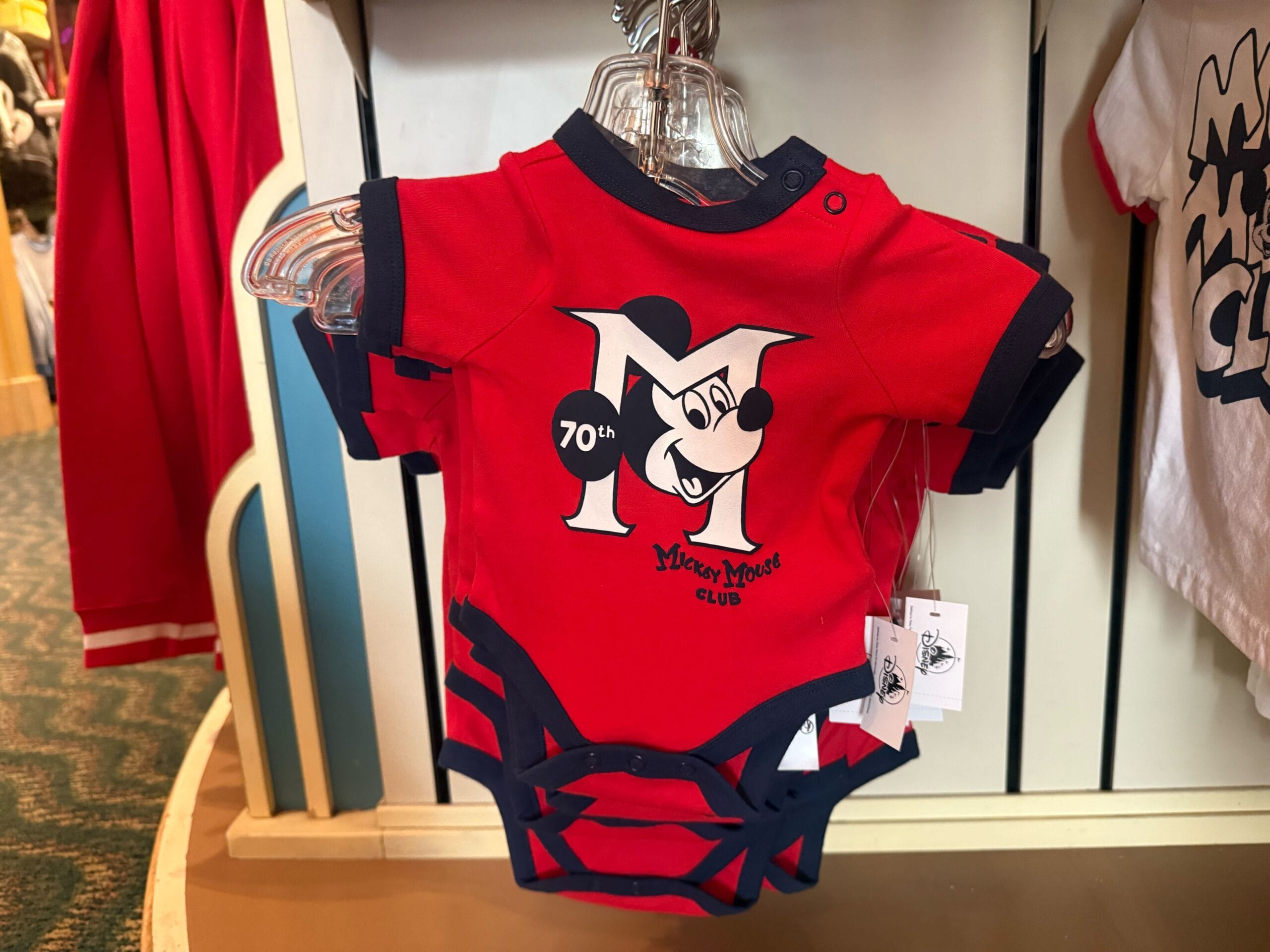 A red baby onesie with "Mickey Mouse Club 70th Anniversary" and Mickey Mouse is displayed on a rack at a Disney park store.