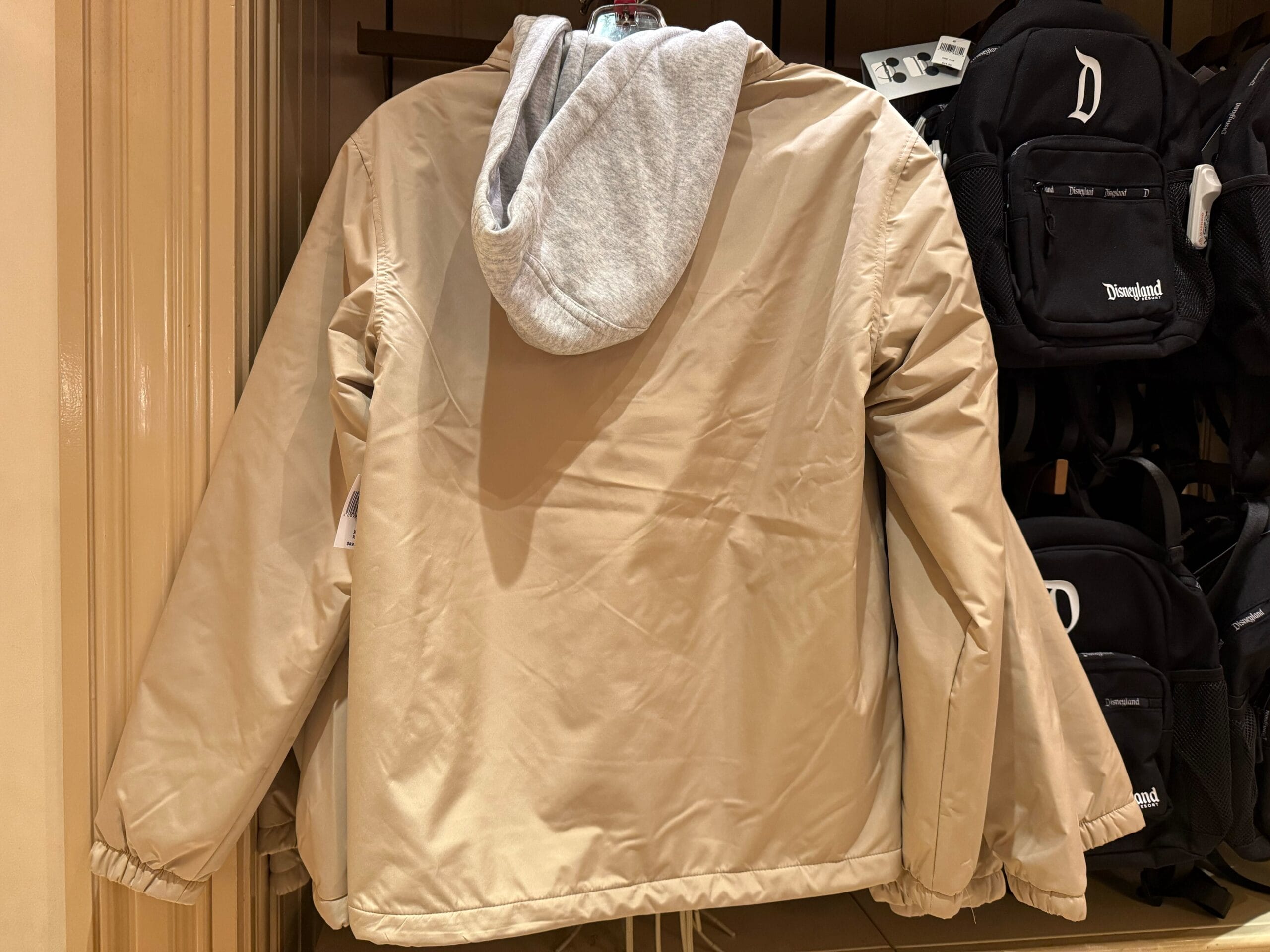 Beige jacket with a gray hood on a rack beside black Disneyland backpacks, likely from a Disney park merchandise store.