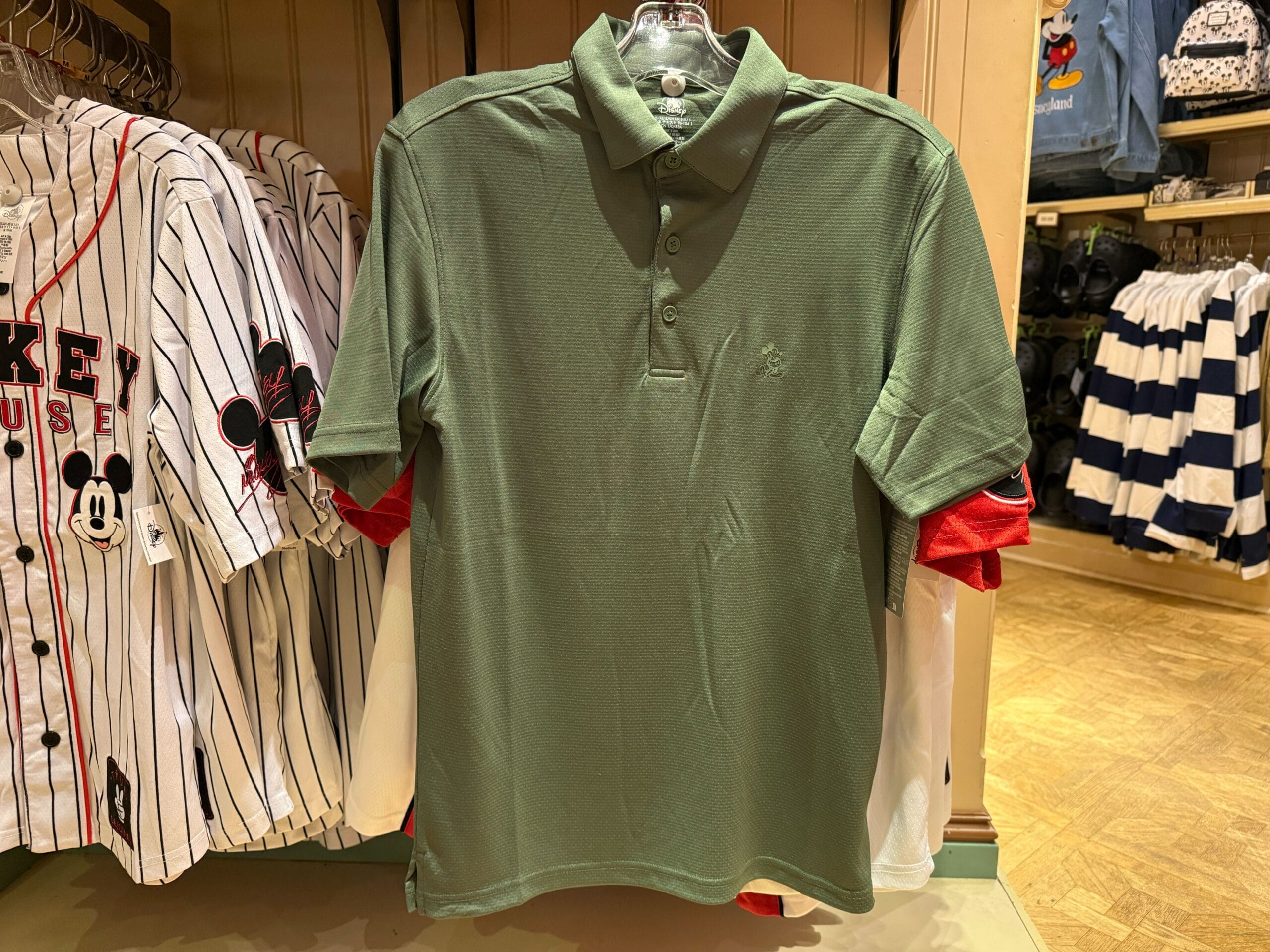A green polo shirt displayed with Mickey Mouse baseball shirts in a Disneyland shop, classic park merch style—no castle in sight.