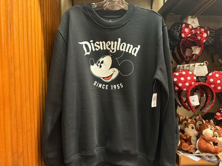 A black Disneyland sweatshirt with Mickey Mouse and "Since 1955," next to Minnie ear headbands, classic Main Street U.S.A. style.
