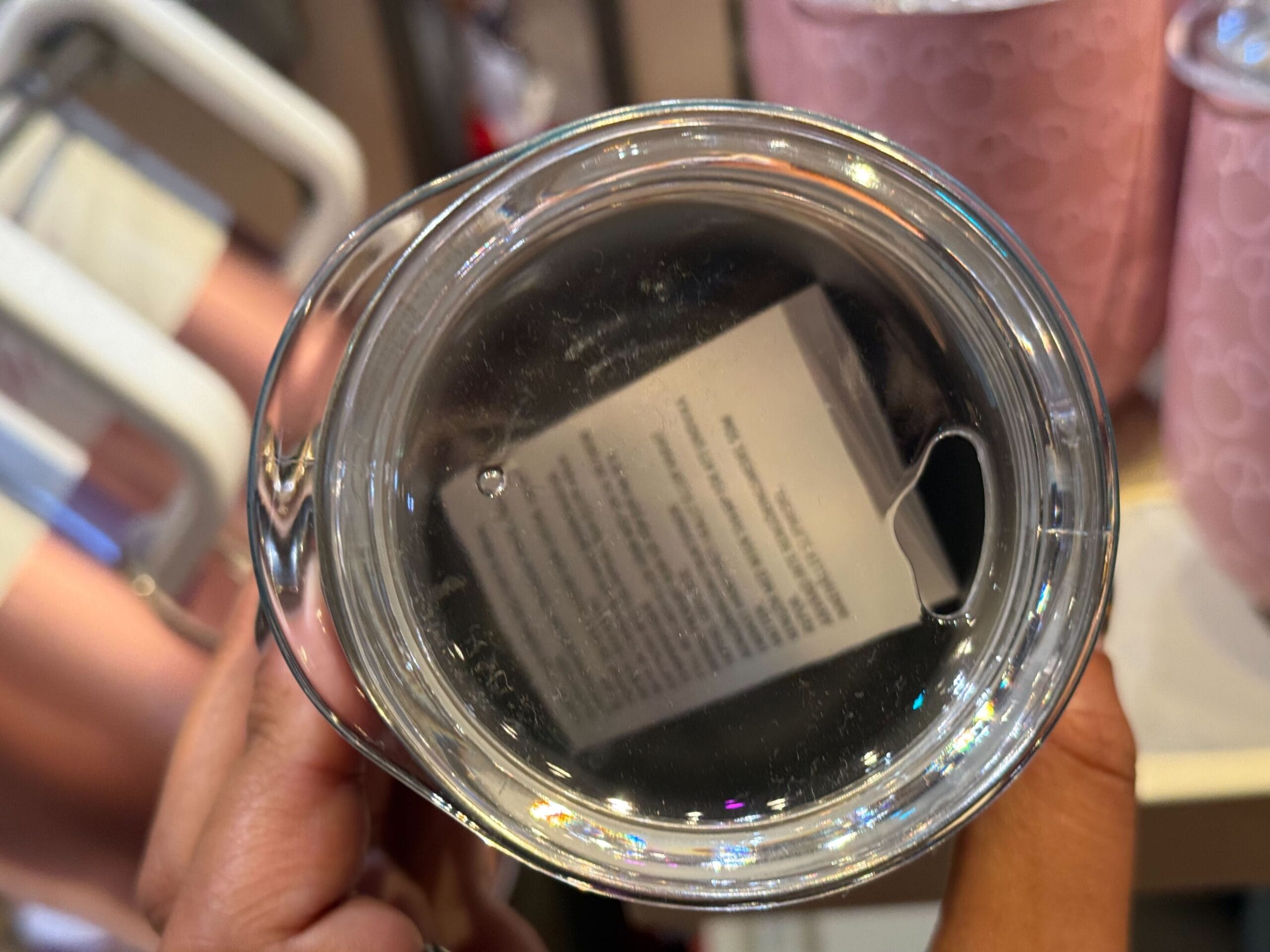 A hand holds a clear glass tumbler with a label and text visible inside, seen from above—much like drinkware sold at Disney parks.