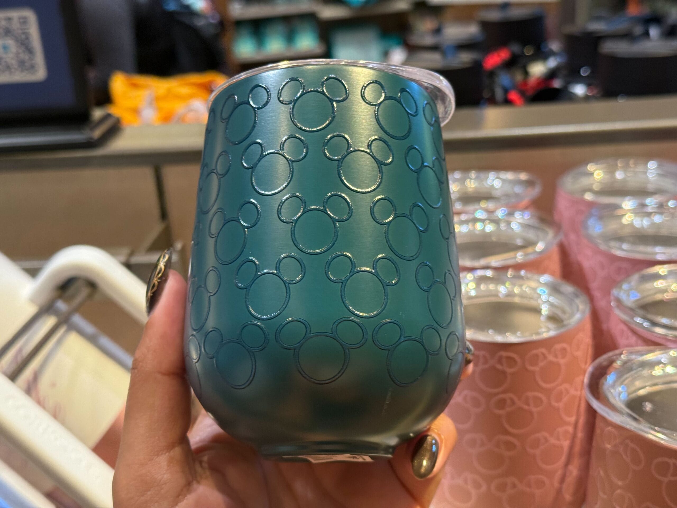 A hand holds a teal metal cup with embossed Mickey heads, reminiscent of Disney Parks merchandise; pink cups sit behind.