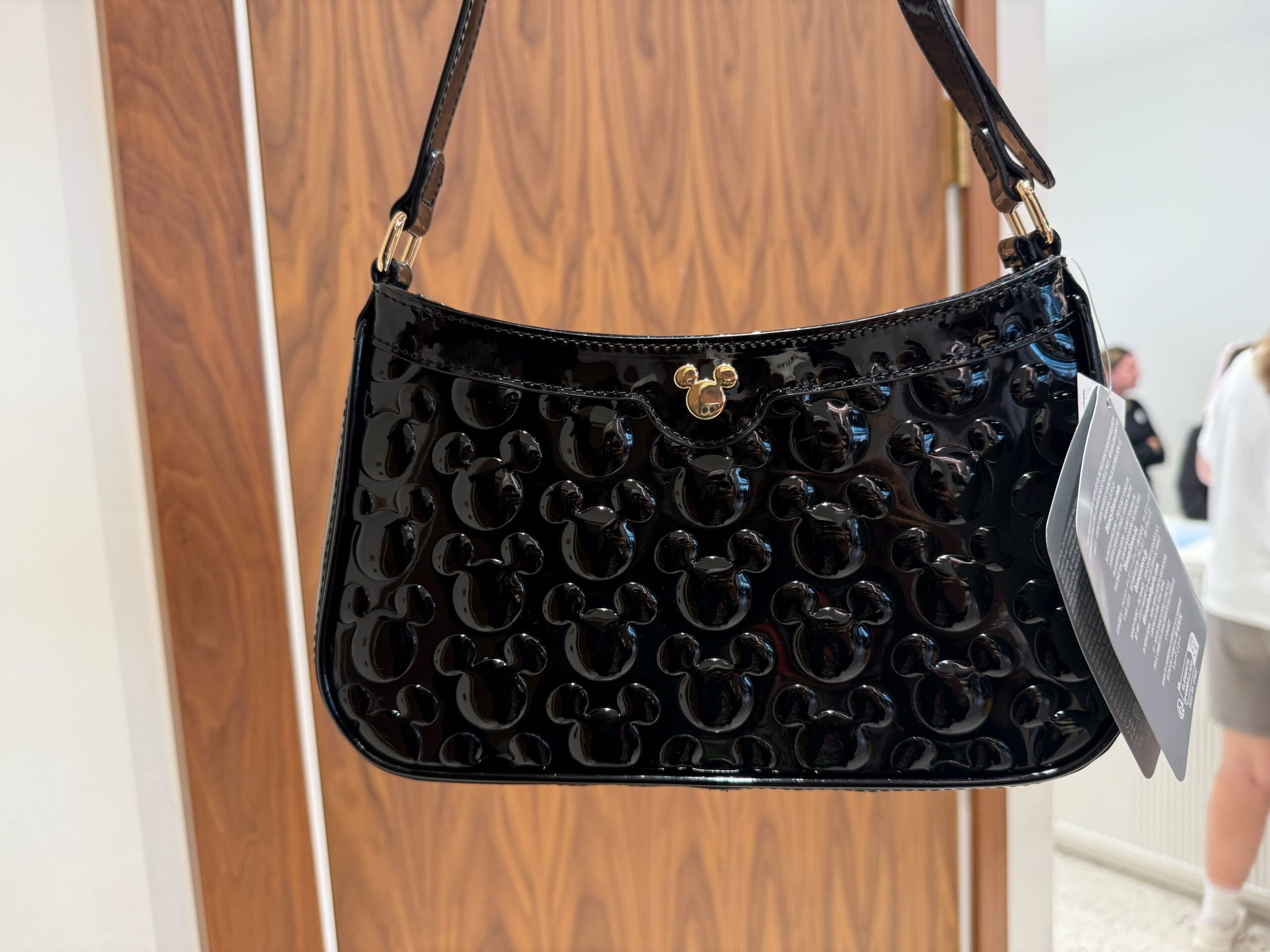 A glossy black purse with embossed Mickey Mouse heads and a gold clasp, hanging before a wooden door in a Disney park setting.
