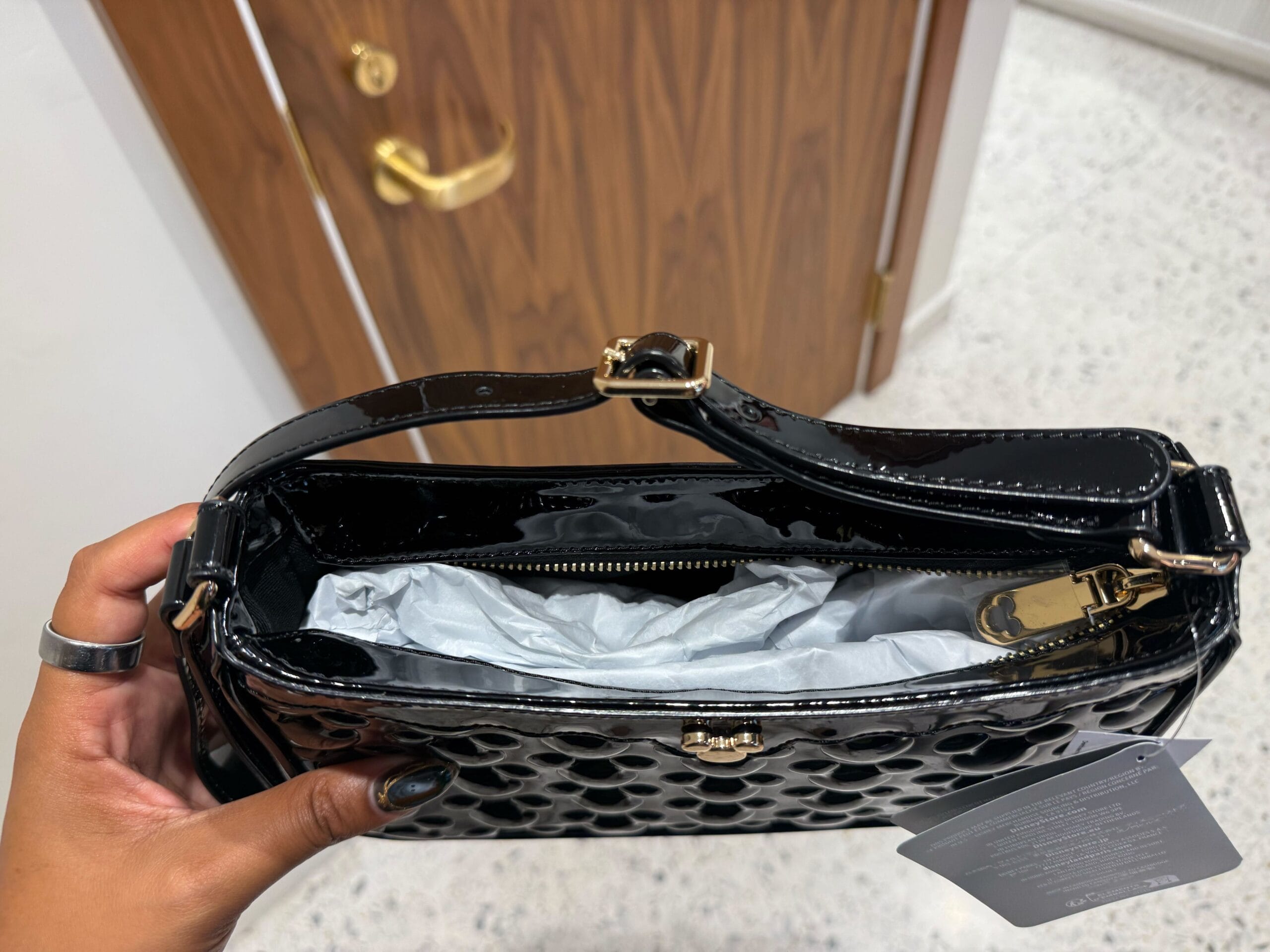 Hand holding a black Icon Purses patent leather purse with gold hardware, opened like opening a Disney park souvenir bag—tissue inside.
