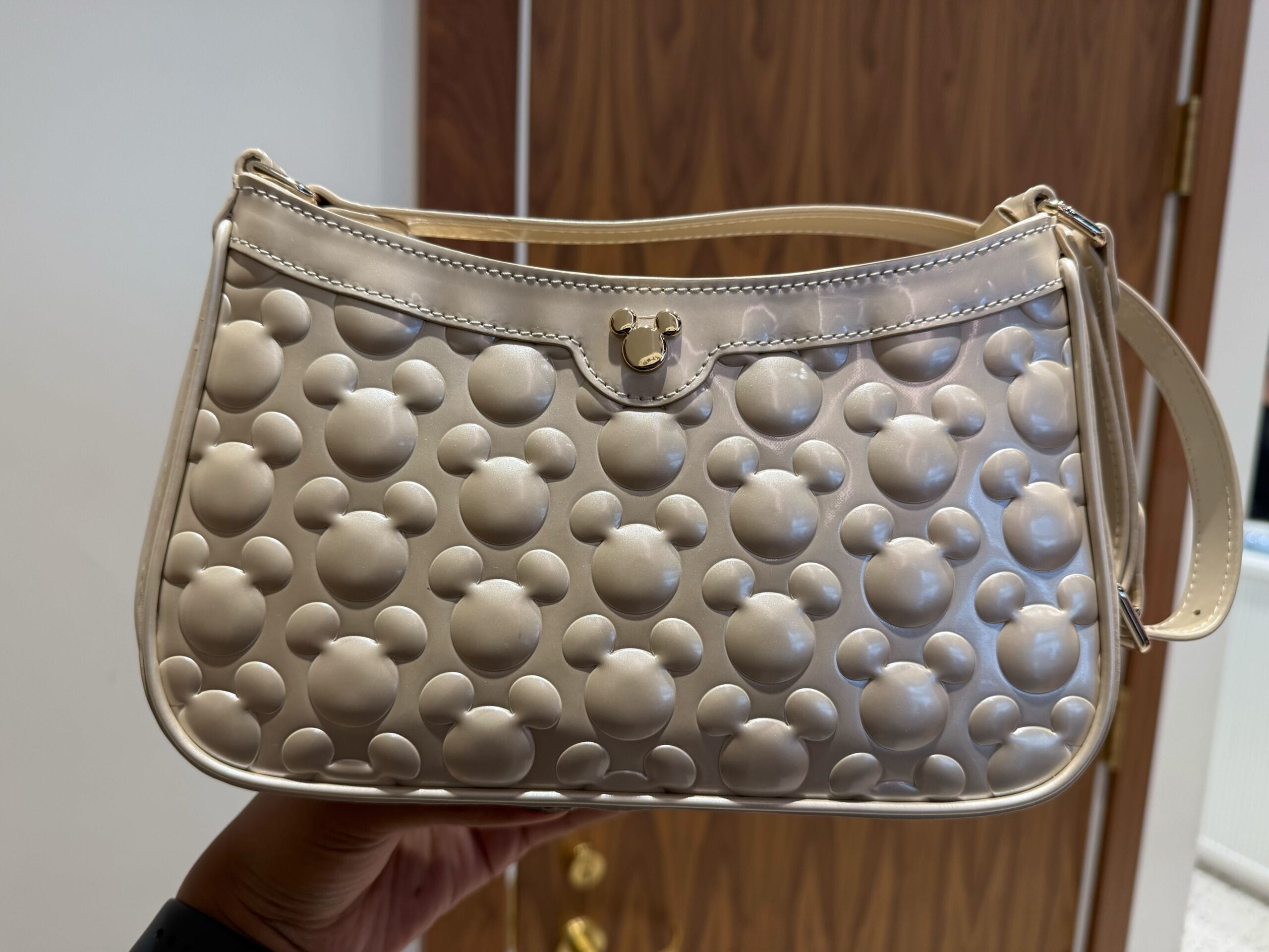 A beige Icon Purse with embossed Mickey Mouse heads, held in front of a wooden door at a Disney Park, no castle in view.