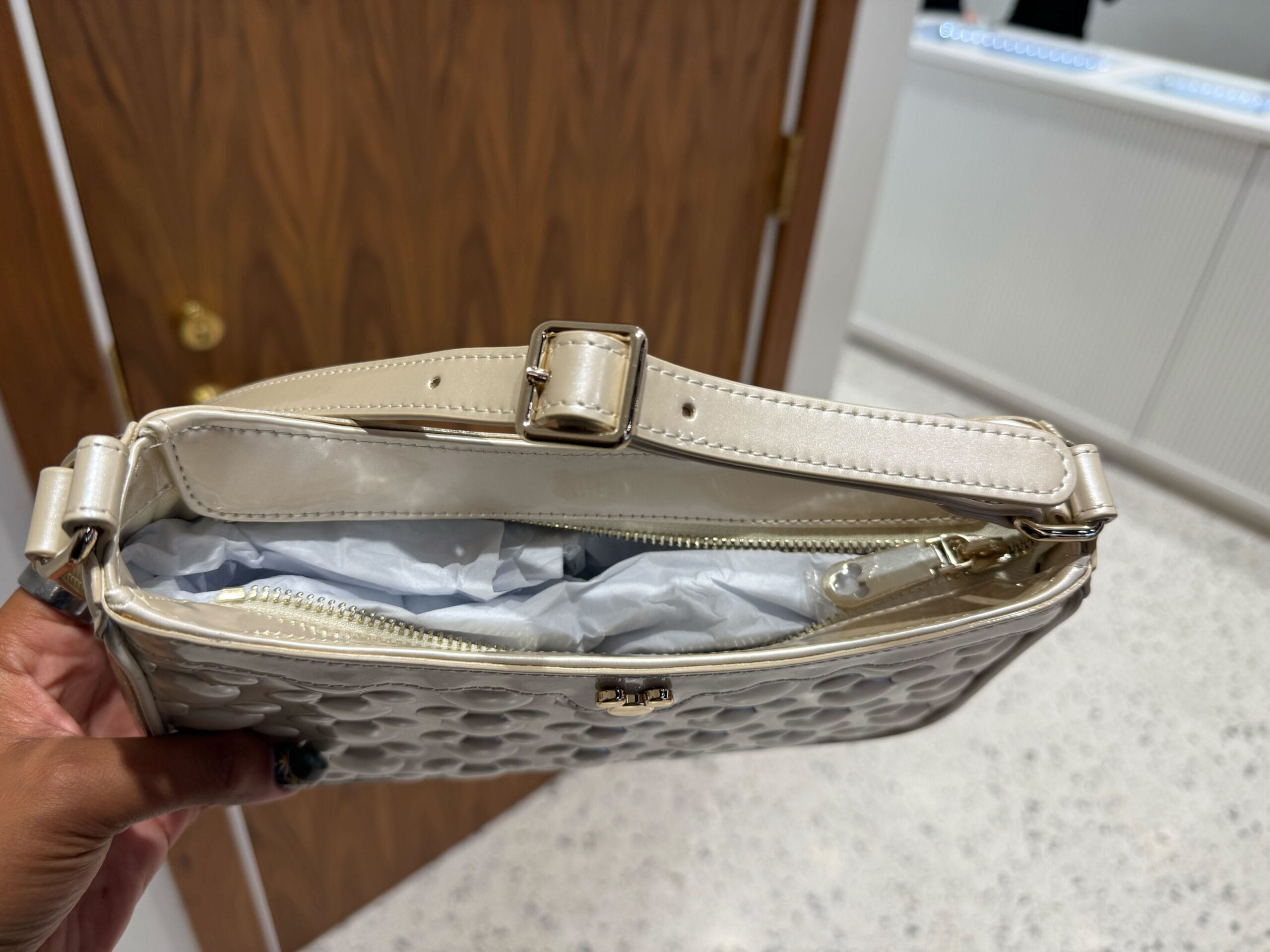A hand holds an open cream-colored Icon Purse filled with tissue paper, near a wooden door—not unlike Main Street shop doors at Disney Parks.