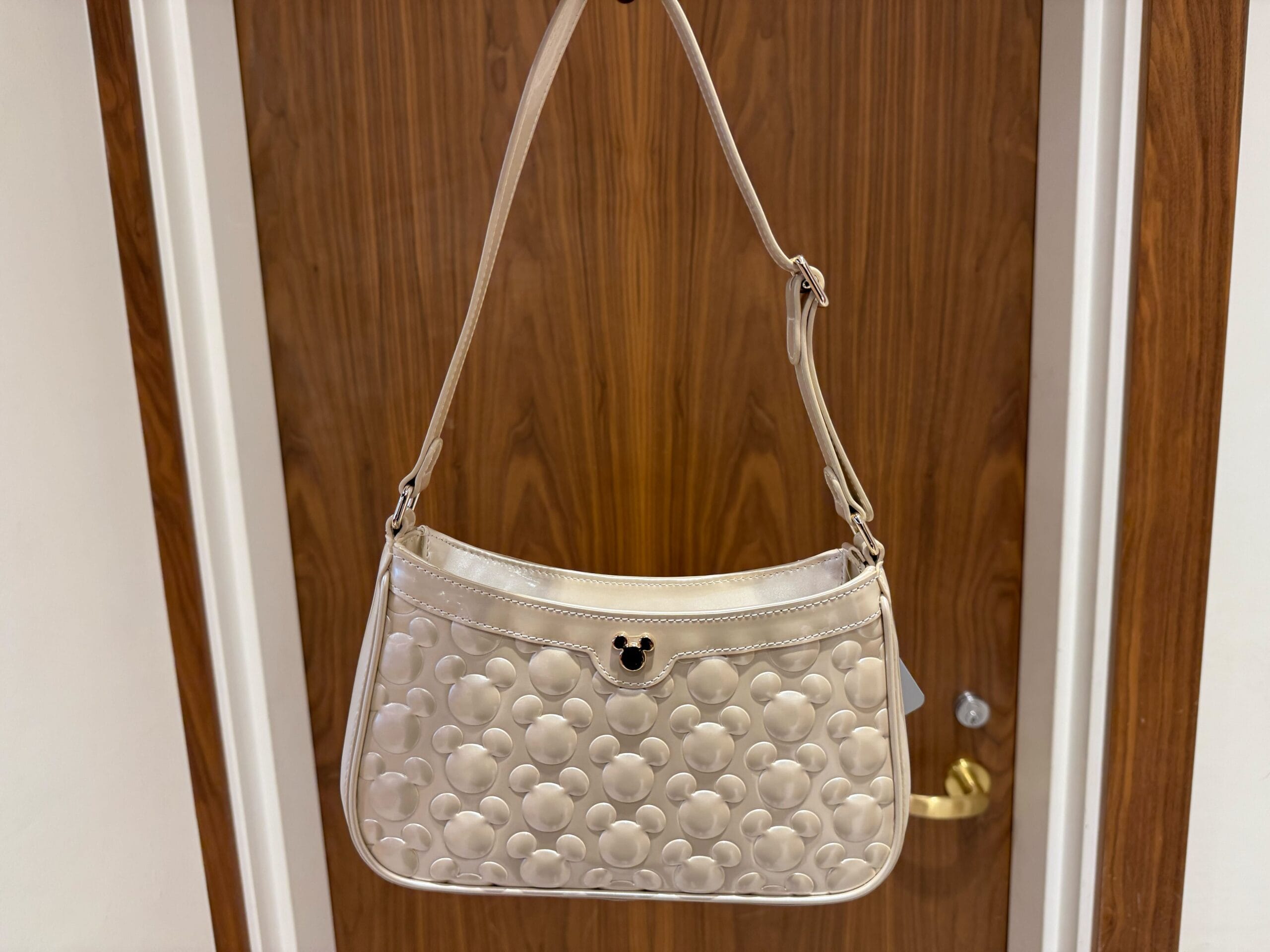 Beige purse with embossed Mickey Mouse pattern, hanging on a wooden door with gold handle, reminiscent of Disney park merchandise.